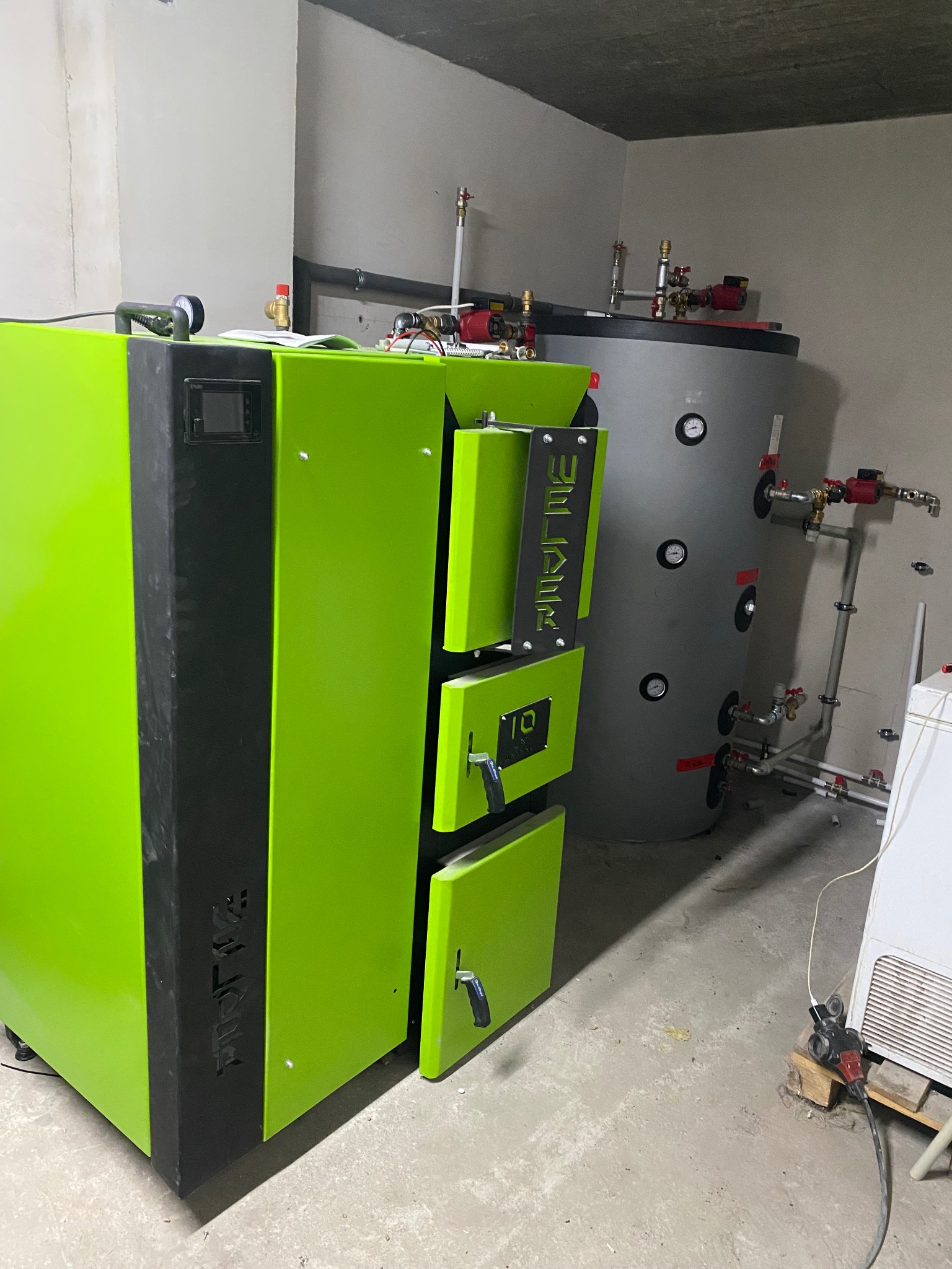 Jasnozielony piec na paliwo stałe z czarnymi akcentami, obok szary zbiornik akumulacyjny z manometrami, w surowym pomieszczeniu z betonową podłogą i sufitem.