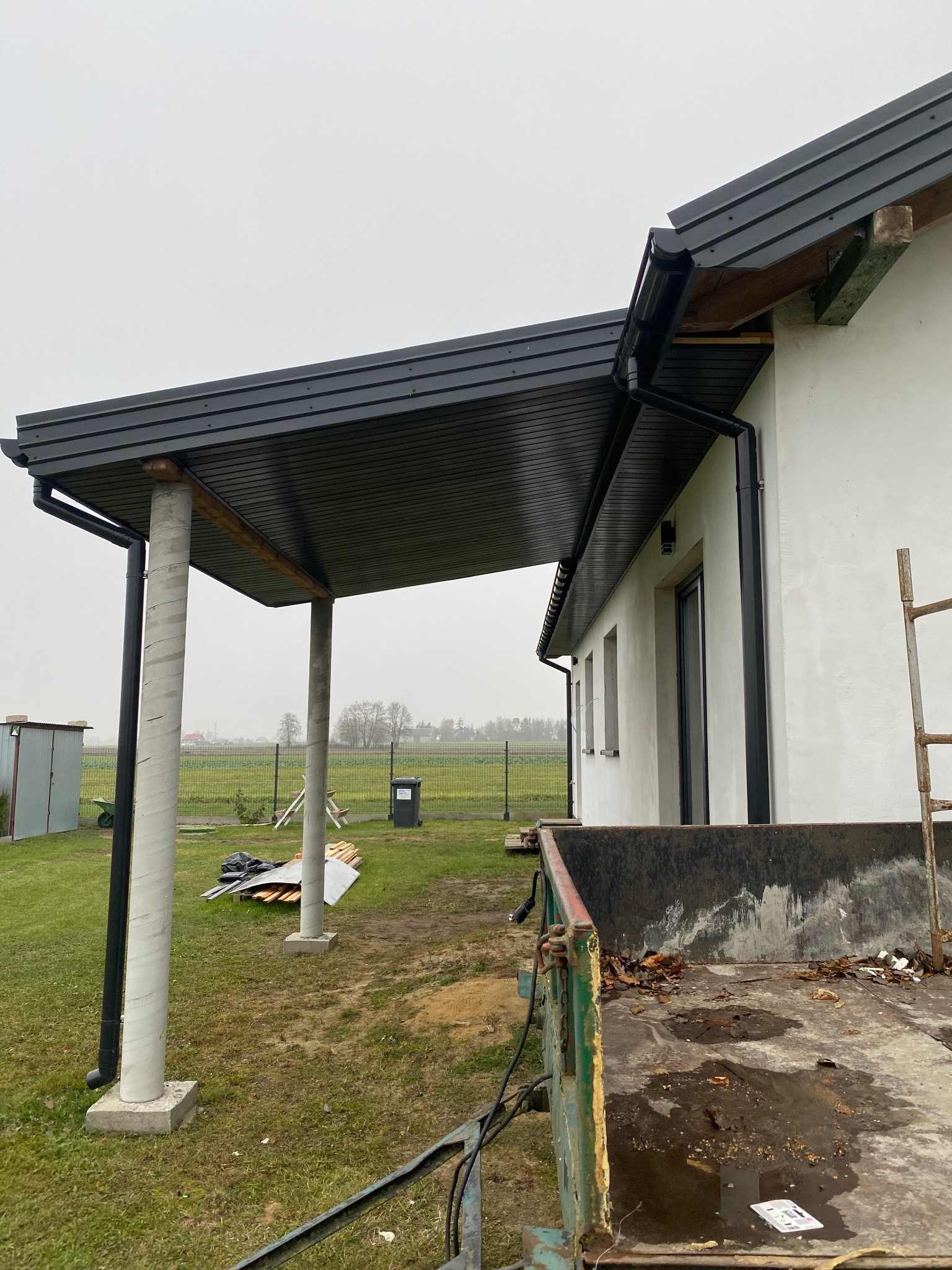 Nowoczesna wiata z grafitowym dachem z blachy trapezowej, wsparta na betonowych kolumnach, widok z boku, z kontenerem na odpady budowlane w pierwszym planie.