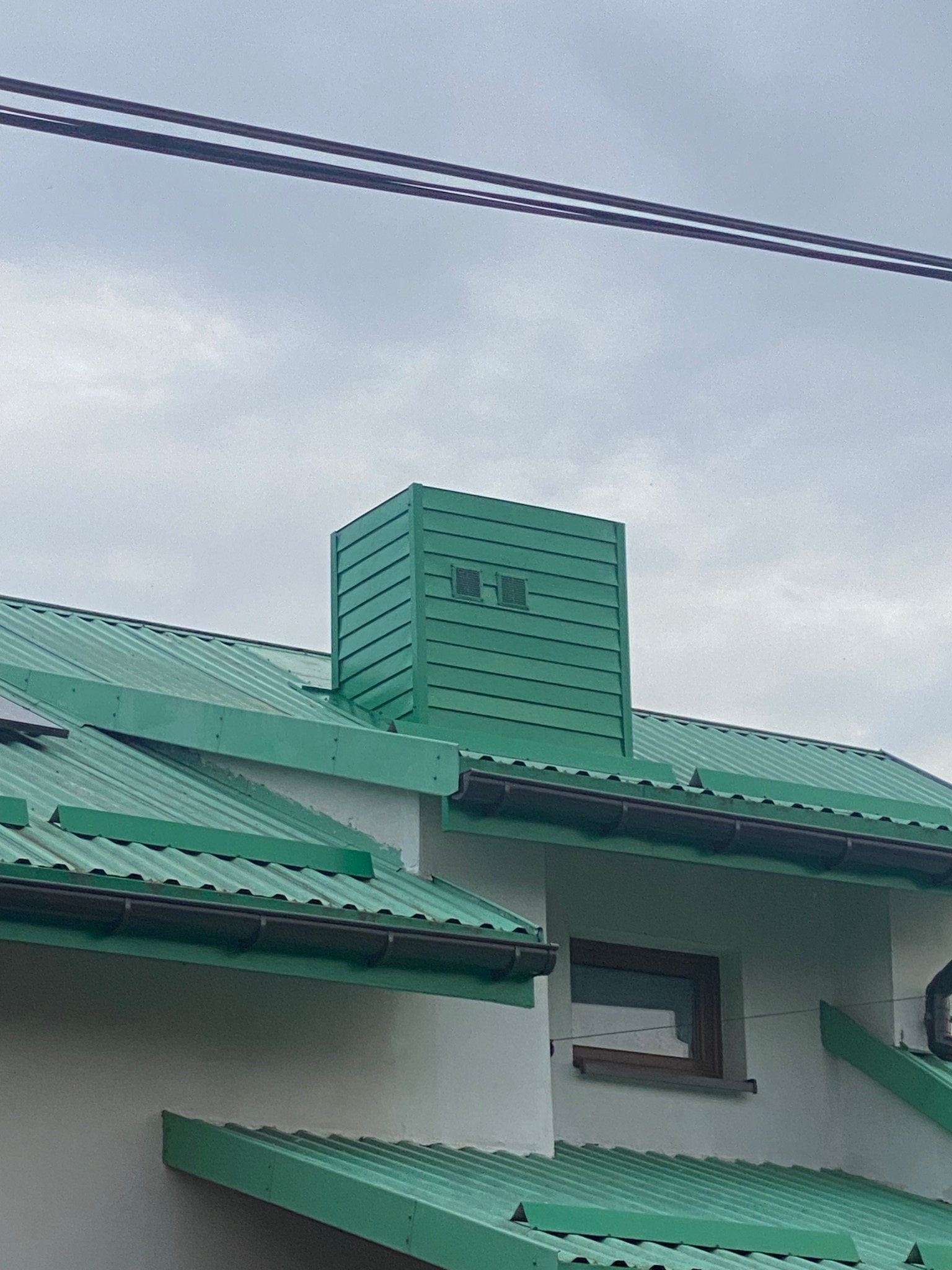 Zielony, blaszany dach z kominem wentylacyjnym i rynnami na tle pochmurnego nieba.