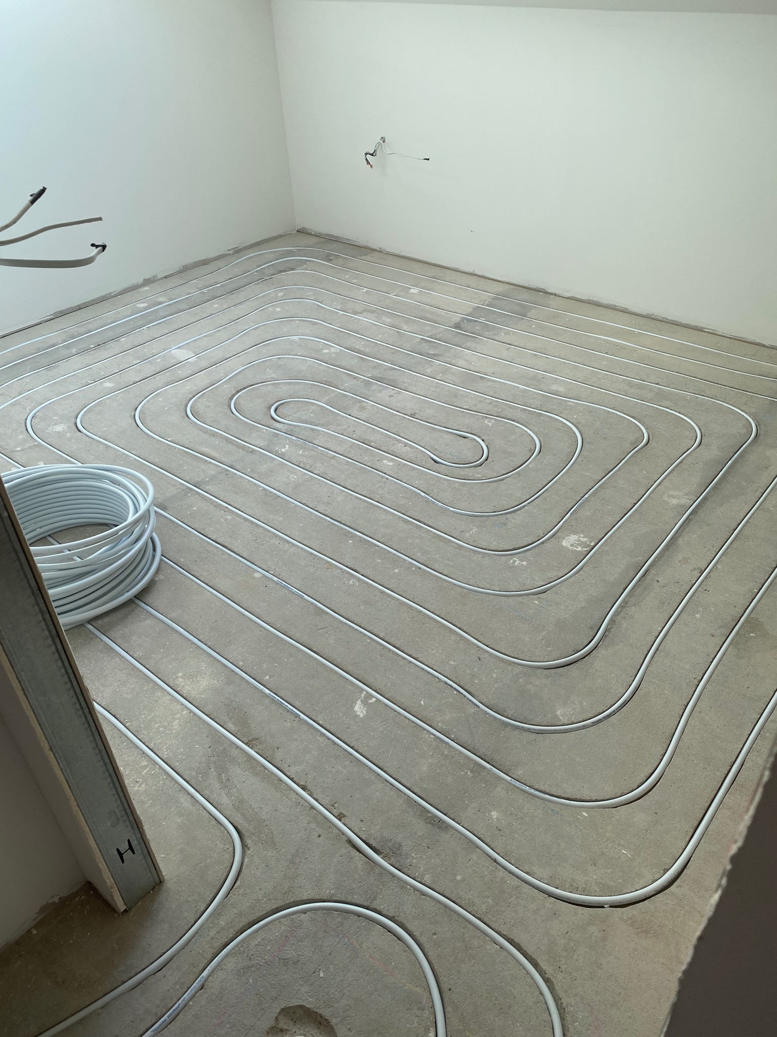 Białe rurki ogrzewania podłogowego rozłożone spiralnie na szarym betonie w trakcie montażu, widoczna rolka rur i fragment ściany z wystającymi przewodami.