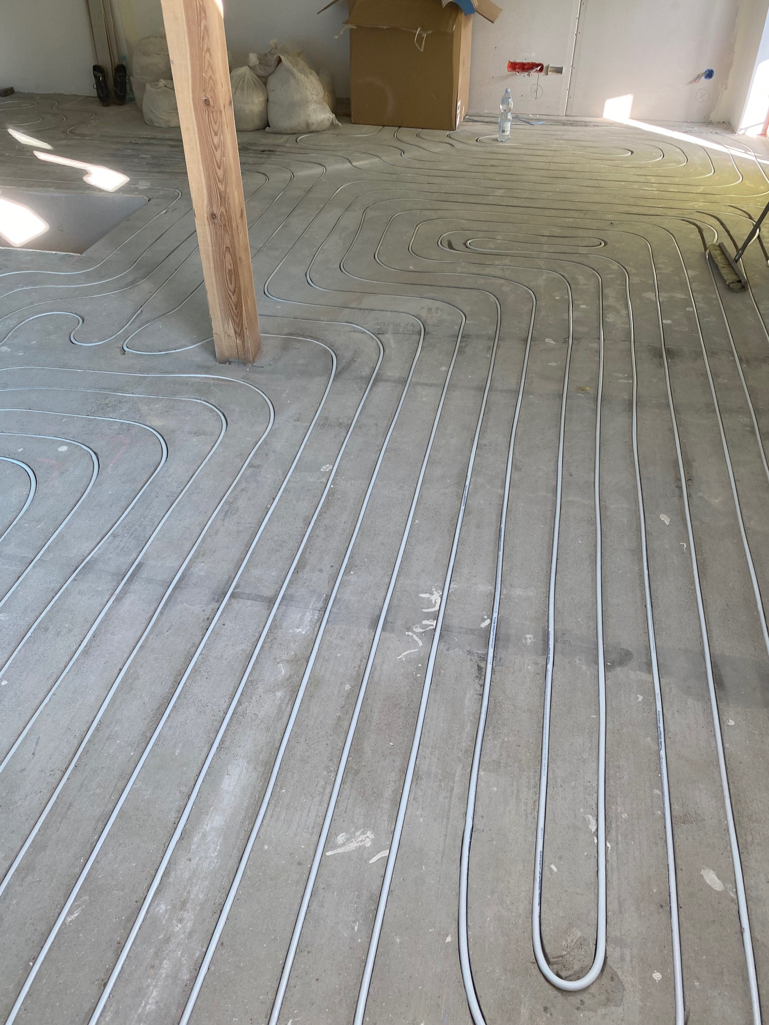 Białe rurki ogrzewania podłogowego ułożone w pętle na szarej, betonowej wylewce w trakcie budowy, widoczny drewniany słup i worki z materiałami budowlanymi w tle.