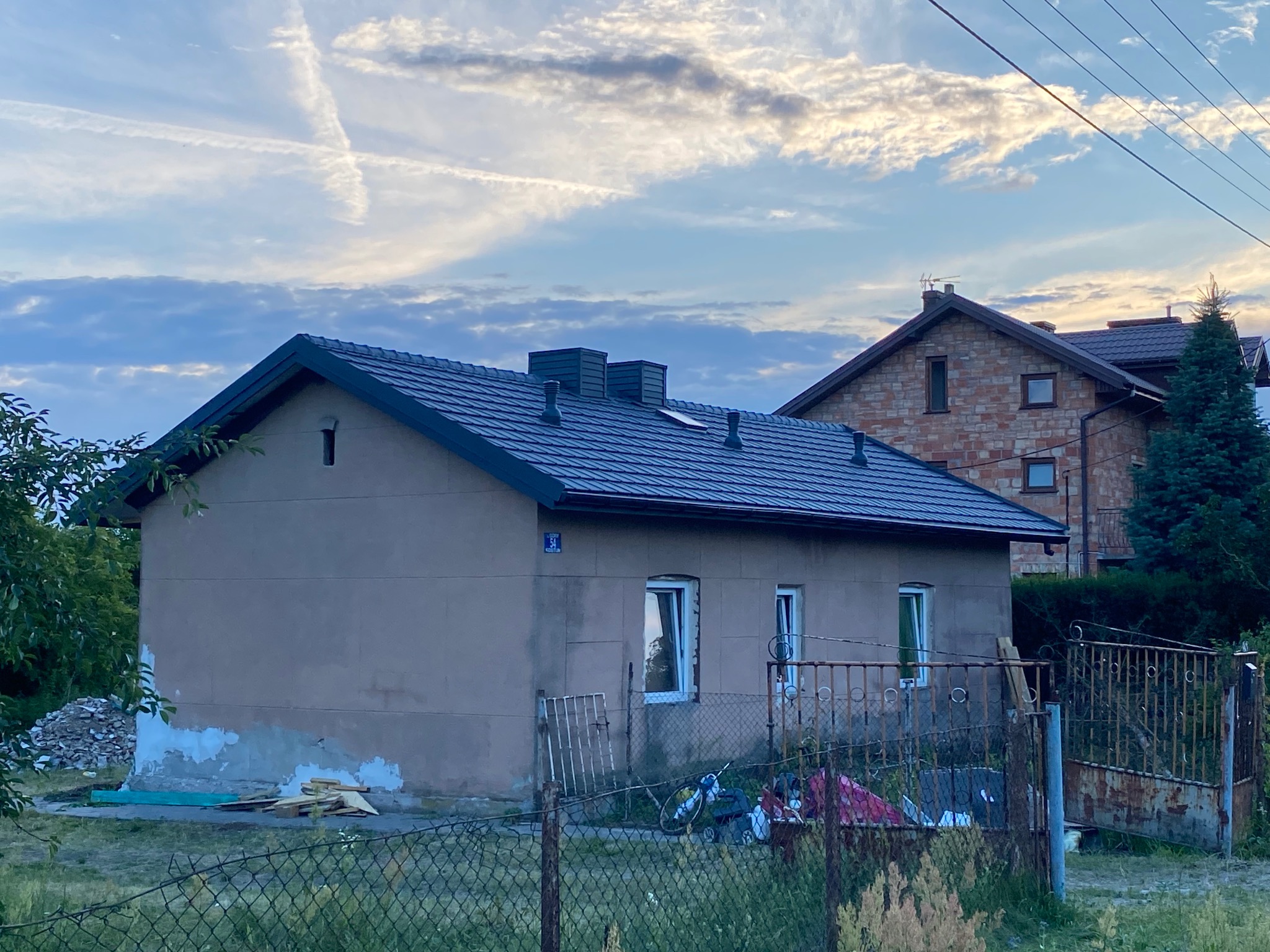 Dom jednorodzinny z nowym, grafitowym dachem z blachodachówki modułowej, widoczne kominy wentylacyjne, w tle drugi dom z cegły, niebo z smugami kondensacyjnymi.