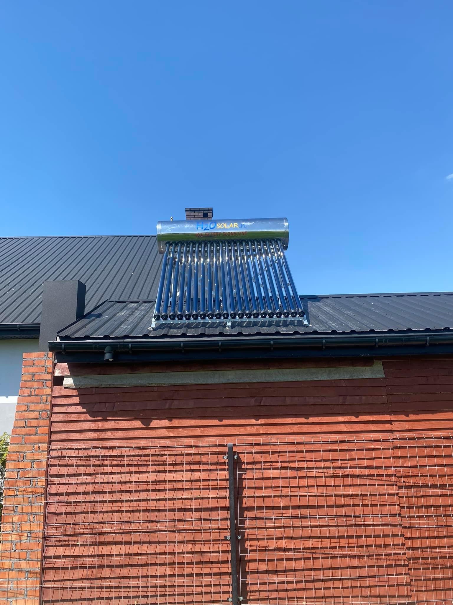 Kolektor słoneczny rurowy zamontowany na dachu z blachy trapezowej, widoczny komin, czerwona drewniana elewacja ściany budynku w tle, błękitne niebo.