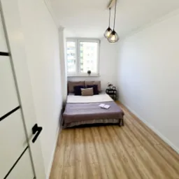 Mała sypialnia z drewnianą podłogą, łóżkiem pod oknem i nowoczesnym oświetleniem, widok na świeżo pomalowane białe ściany i listwy przypodłogowe.