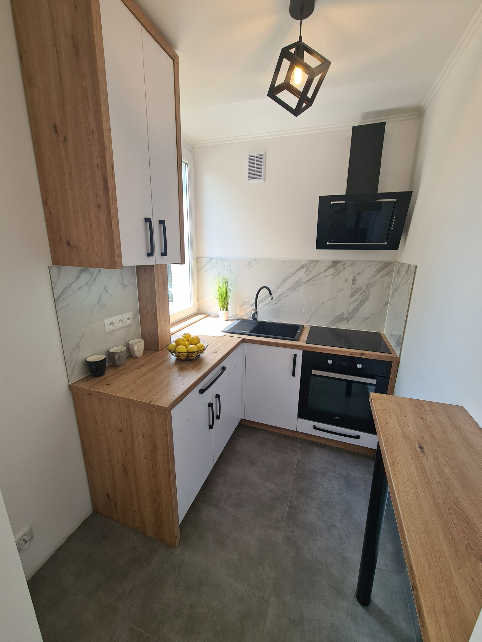 Nowoczesna, kompaktowa kuchnia w bieli i drewnie z czarnymi akcentami: okap, zlew, piekarnik, lampa sufitowa. Na blacie misa z cytrynami i kilka kubków. Marmurowe panele ścienne.