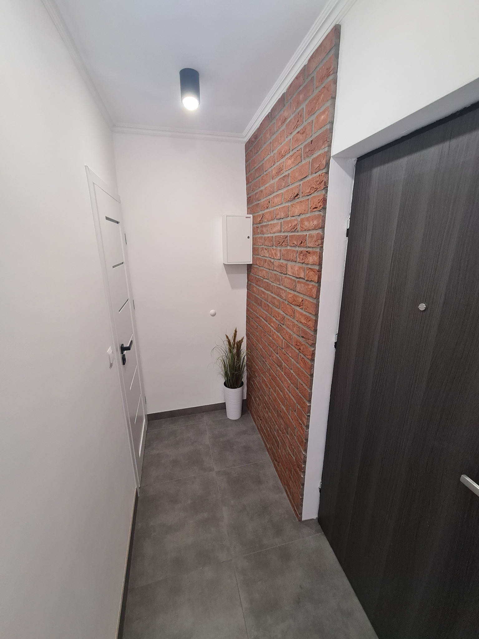Wąski korytarz z dekoracyjną ścianą z czerwonej cegły, szarą podłogą z płytek, białą szafką elektryczną i wysoką, ciemną, nowoczesną stolarką drzwiową.