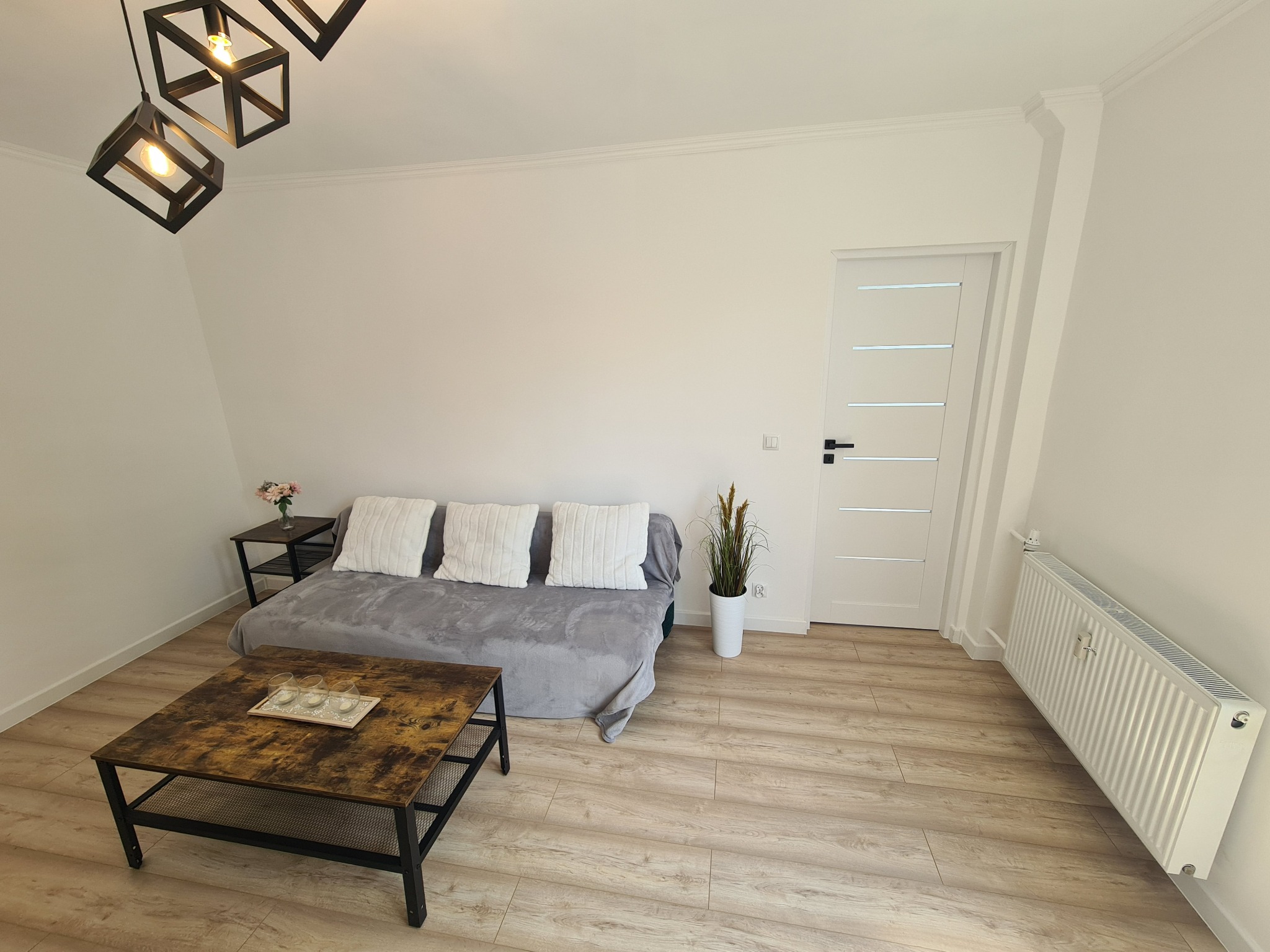 Jasne wnętrze pokoju z drewnianą podłogą, sofą przykrytą szarym pledem z białymi poduszkami, stolikiem kawowym z trzema szklankami, białymi ścianami z listwami przypodłogowymi, białymi drzwiami...