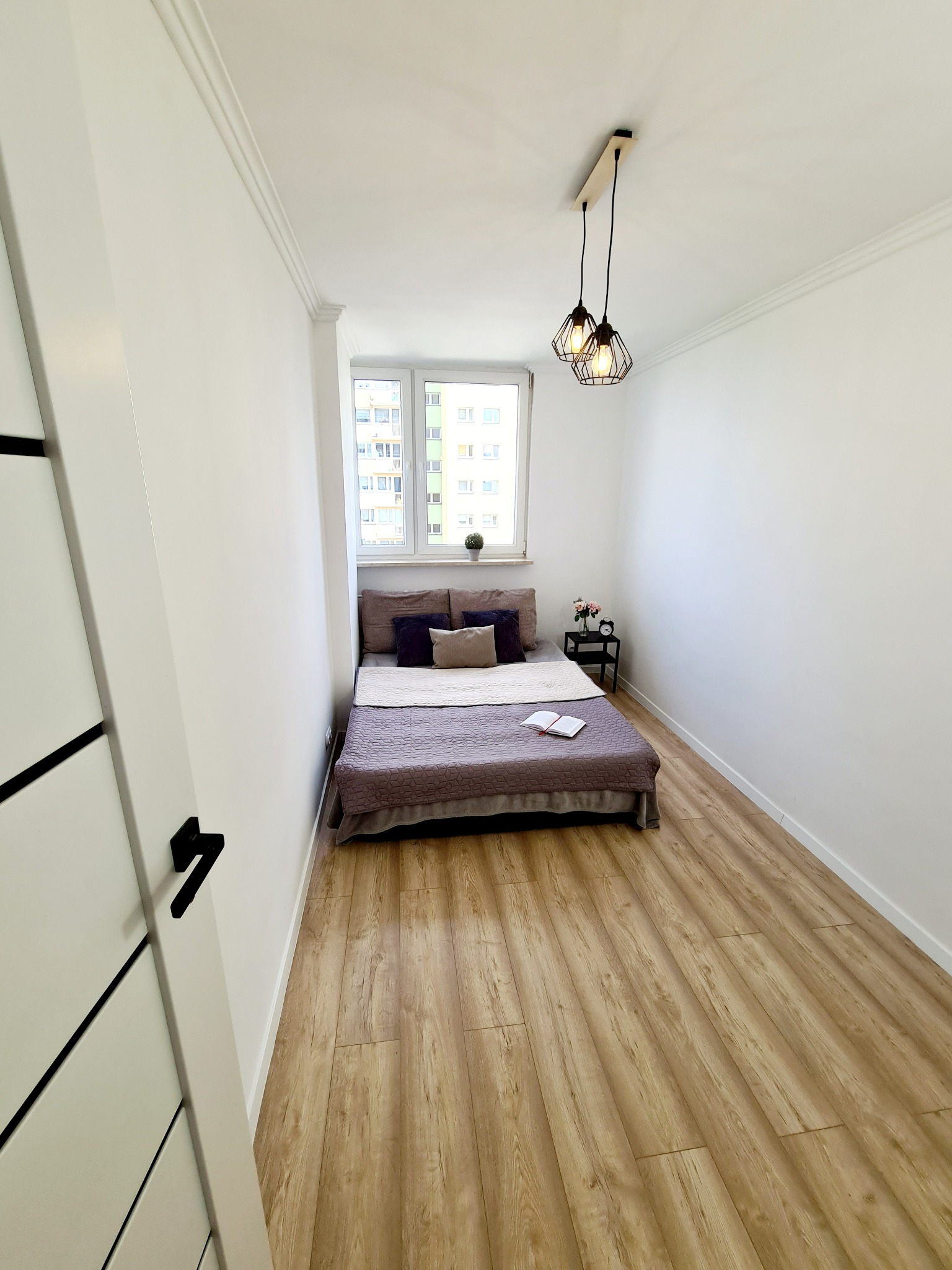 Mała sypialnia z drewnianą podłogą, łóżkiem pod oknem i nowoczesnym oświetleniem, widok na świeżo pomalowane białe ściany i listwy przypodłogowe.