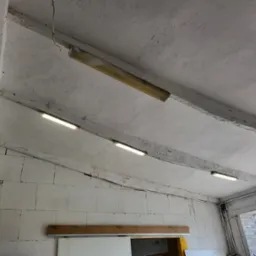 Zamontowane lampy jarzeniowe na betonowym suficie w surowym wnętrzu. Widoczne przewody elektryczne i ściany z białych bloczków. Oświetlenie hali lub magazynu.