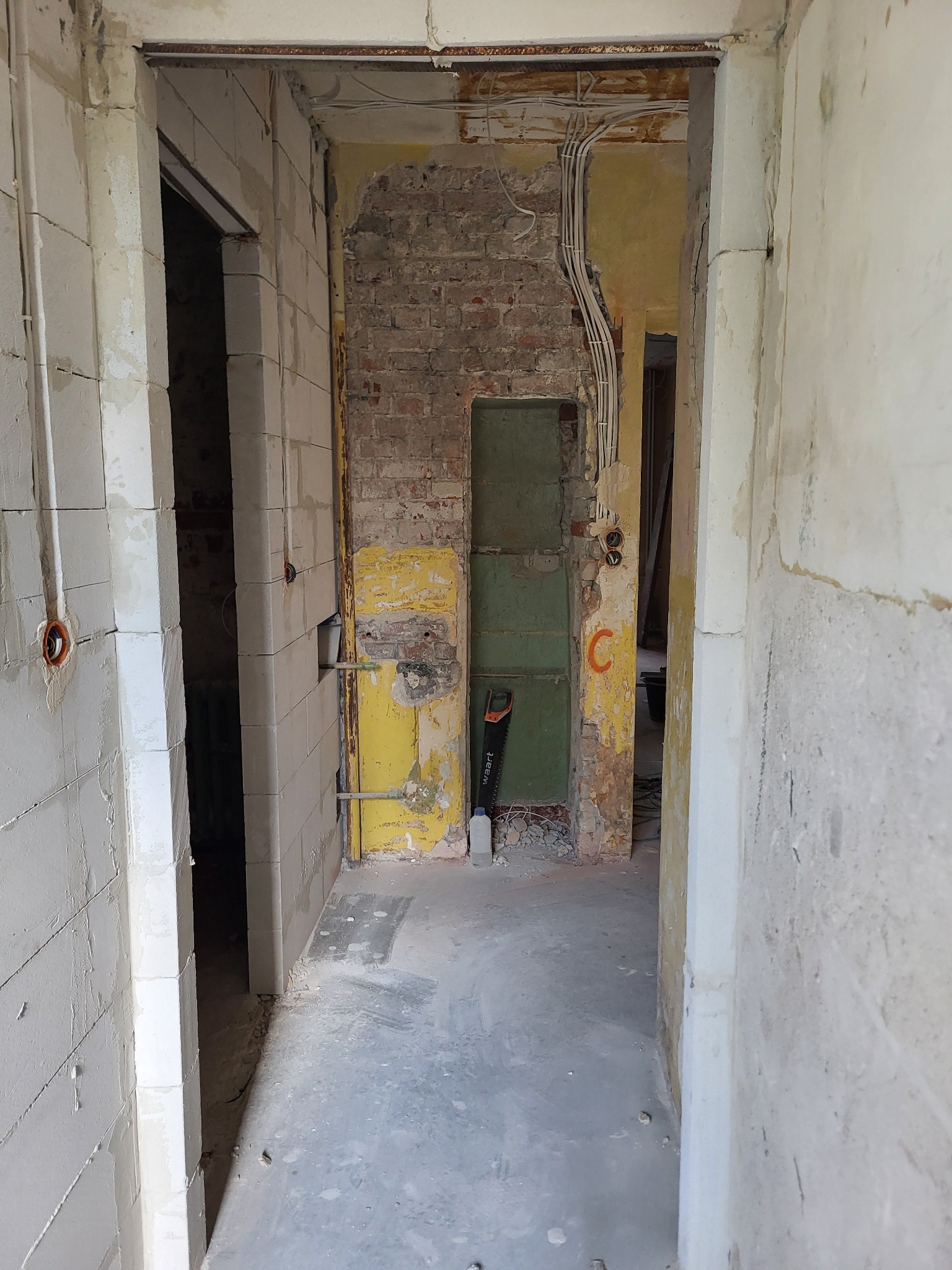 Wnętrze w trakcie remontu: widoczne przewody elektryczne na ścianie z cegły, surowe ściany z bloków i resztki starej farby. Piła leży przy ścianie.