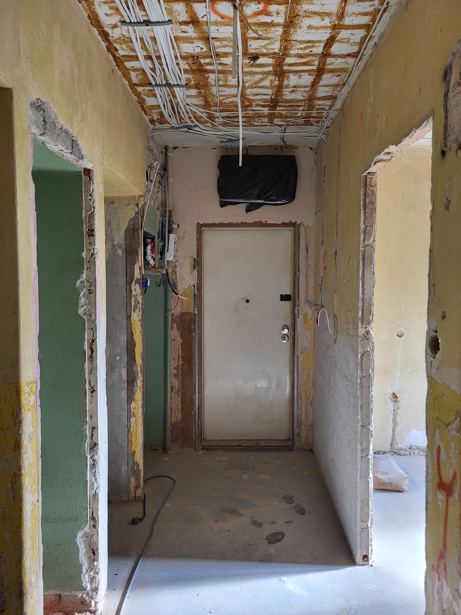 Remont korytarza: widoczne kable na suficie, ściany w trakcie skuwania, surowy beton, przygotowanie do dalszych prac wykończeniowych.