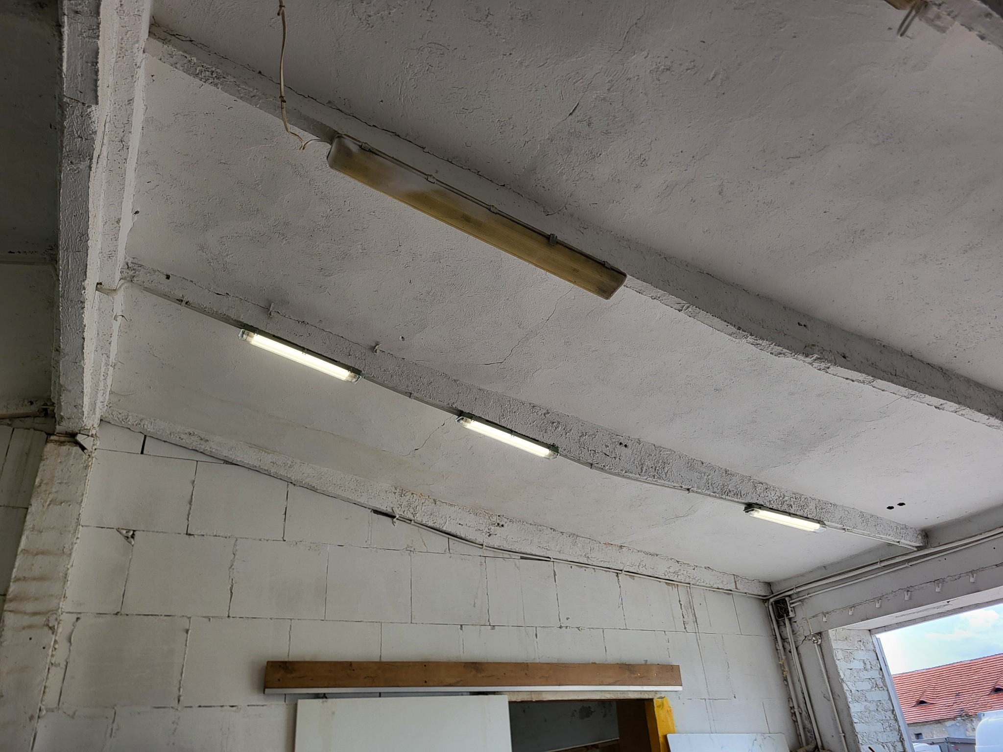 Zamontowane lampy jarzeniowe na betonowym suficie w surowym wnętrzu. Widoczne przewody elektryczne i ściany z białych bloczków. Oświetlenie hali lub magazynu.