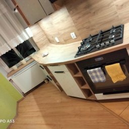 PHOENIX - Kuchnia na wymiar z drewnianym blatem, czarną płytą gazową i piekarnikiem z wiszącymi ściereczkami, widok na okno z roślinami doniczkowymi.