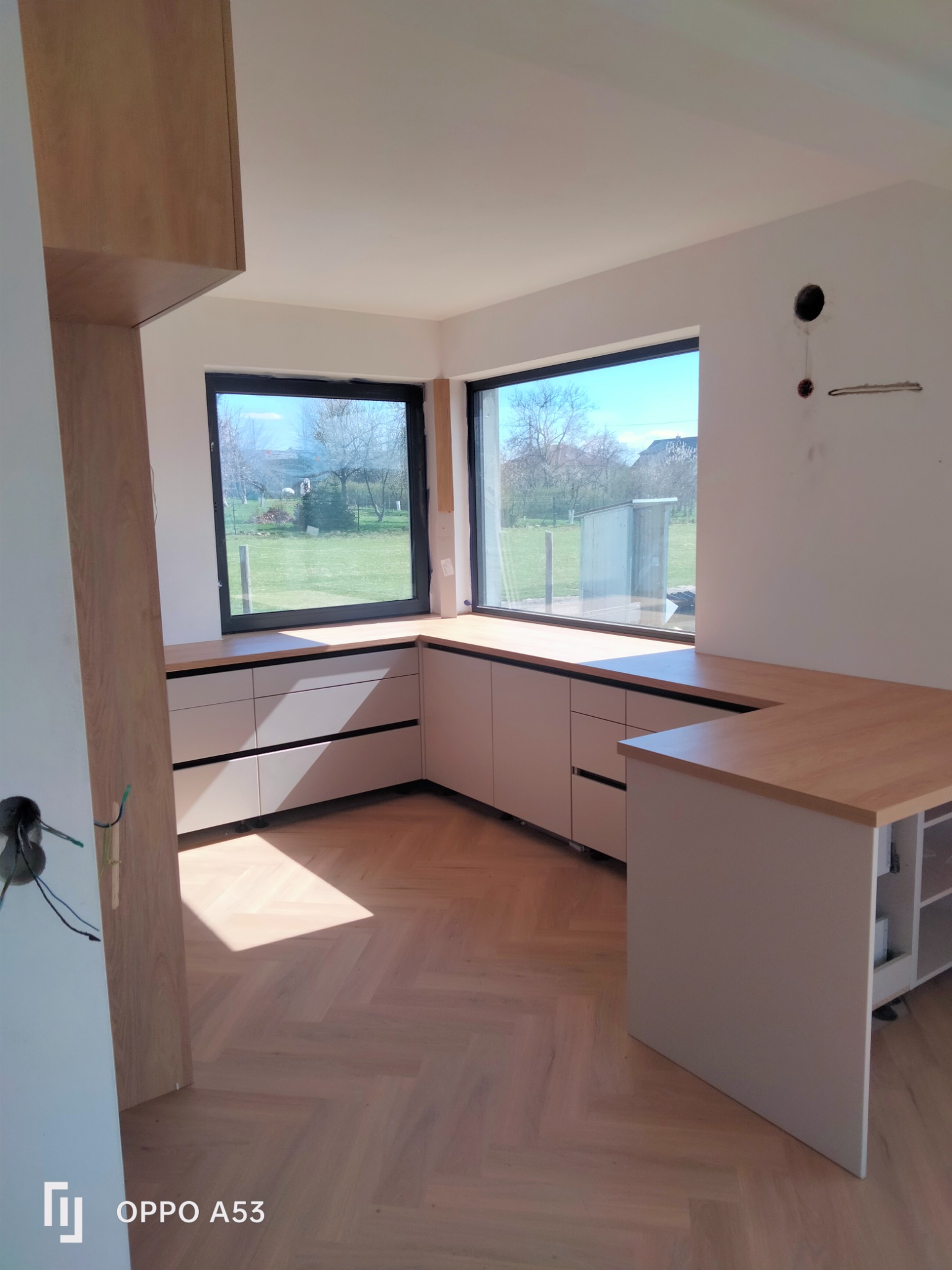 Jasna kuchnia z drewnianym blatem i szufladami, widok na zewnątrz przez okno, parkiet na podłodze, instalacja elektryczna w trakcie.