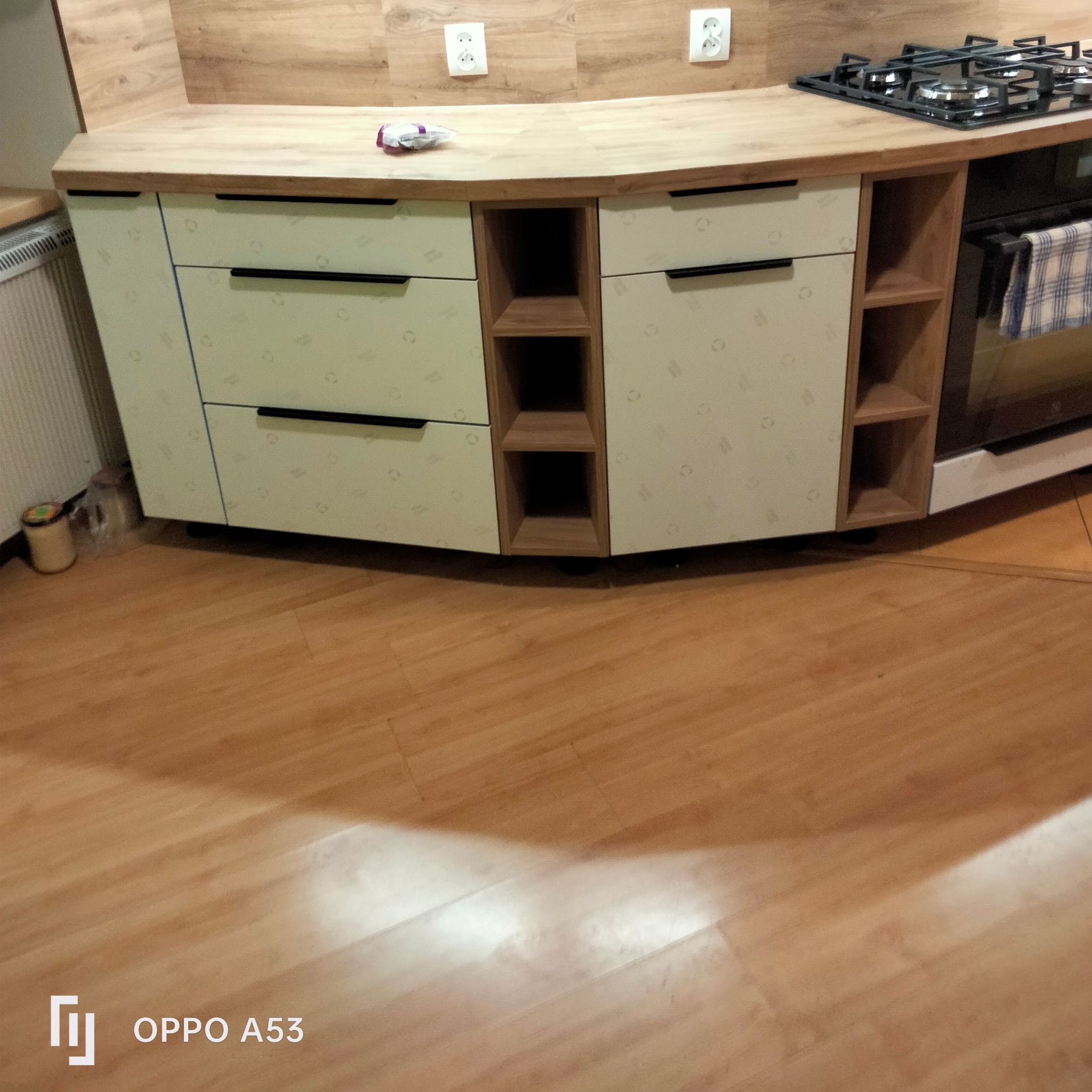 Zaokrąglona zabudowa kuchenna z jasnymi frontami szuflad z delikatnym wzorem, ciemnymi uchwytami, drewnianymi otwartymi półkami, płytą grzewczą i piekarnikiem, widok na drewnianą podłogę i gniazdka...