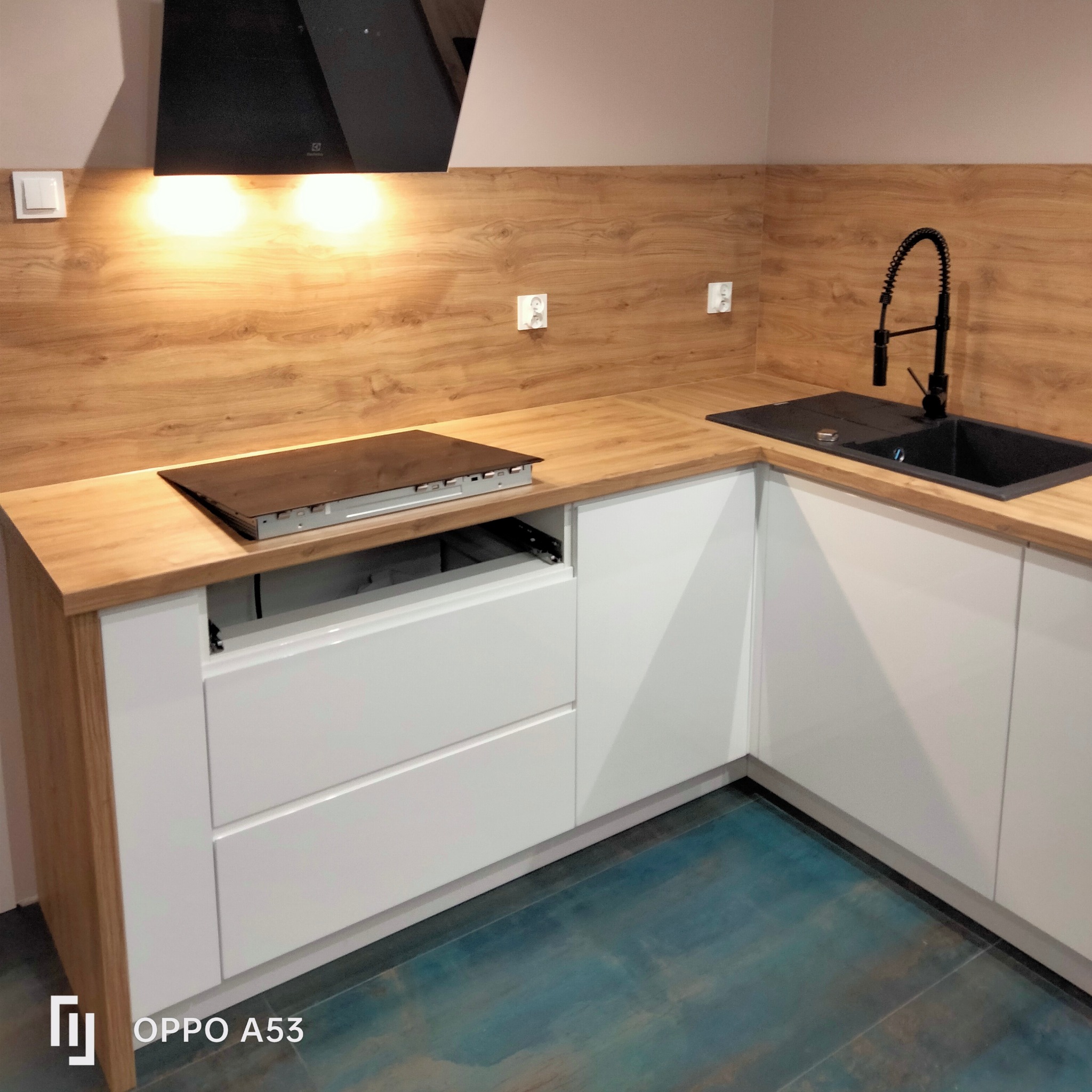 Fragment narożnej zabudowy kuchennej z białymi, lakierowanymi frontami, drewnianym blatem i czarnym zlewem z baterią, widoczna płyta indukcyjna z wysuniętym szufladami. Na ścianie drewnopodobna...