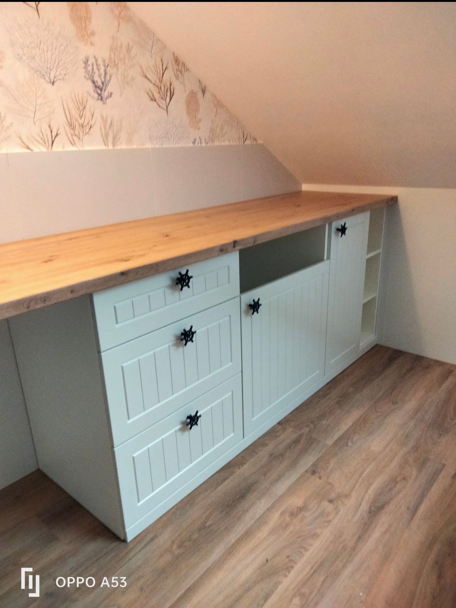 Biała zabudowa meblowa pod skosem z drewnianym blatem i dekoracyjnymi, czarnymi uchwytami w kształcie gwiazd, na podłodze panele drewnopodobne, ściana pokryta tapetą w roślinne wzory.