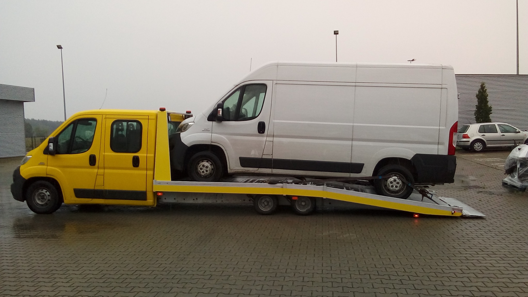 Żółta autolaweta z białym busem na platformie, widok z boku na mokrym bruku, w tle budynek z blachy falistej i zaparkowane auto.