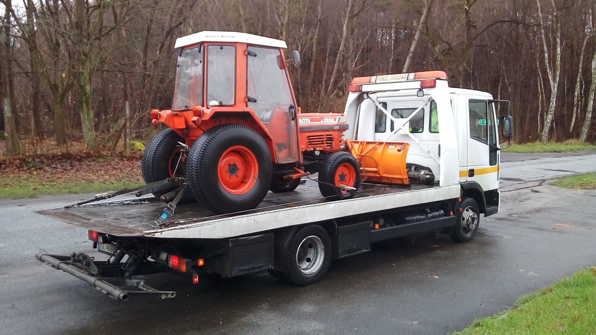 Pomarańczowy traktor rolniczy z pługiem śnieżnym przewożony na białej platformie pojazdu pomocy drogowej na tle drzew bez liści.
