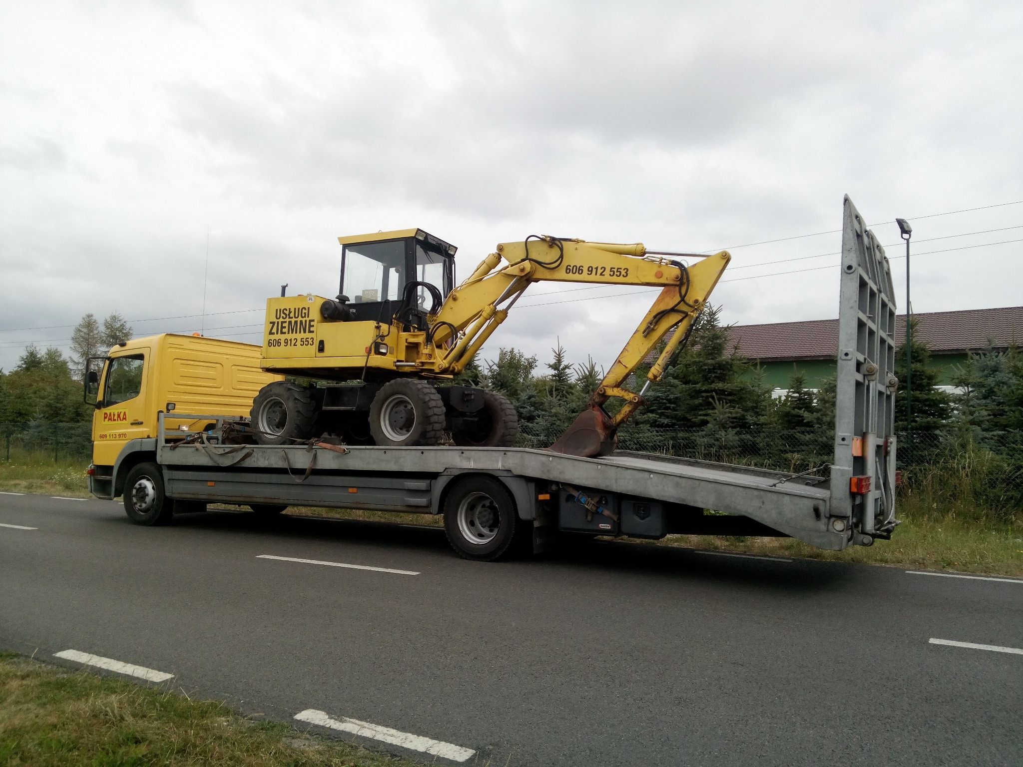 Żółta koparka kołowa załadowana na niskopodwoziową lawetę transportową, widok z boku na asfaltowej drodze, w tle zieleń i budynek.