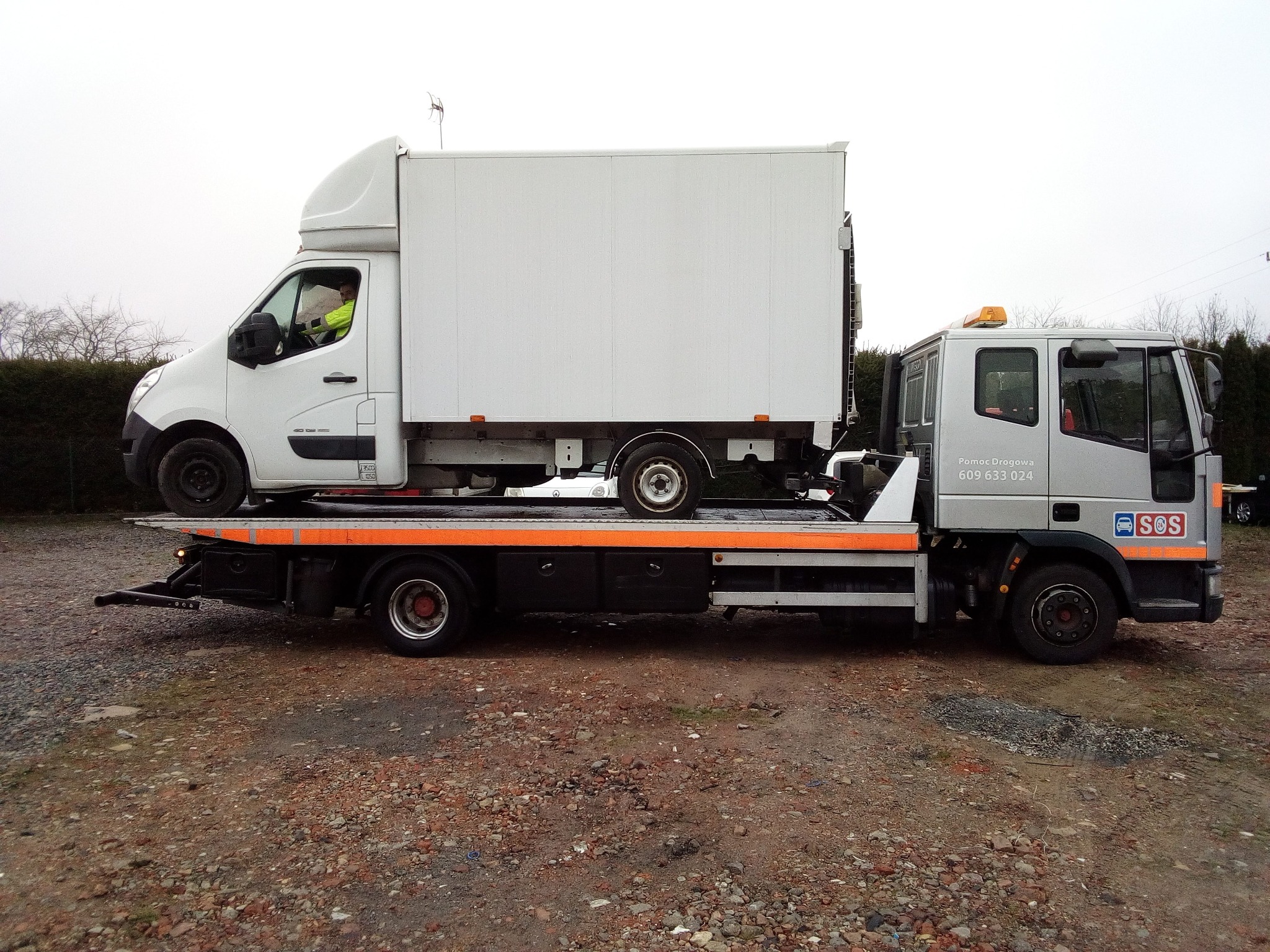 Laweta przewozi biały samochód dostawczy na platformie transportowej z widocznym kierowcą w żółtej kamizelce odblaskowej, na tle szarego nieba i zielonego żywopłotu.