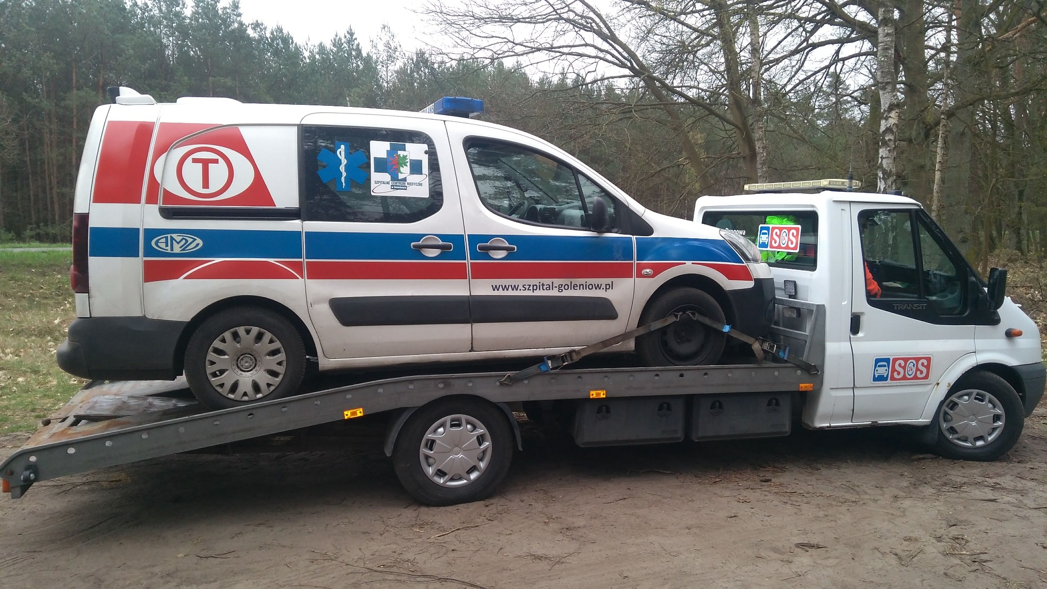 Laweta przewozi ambulans z oznaczeniami szpitala, zabezpieczony pasami transportowymi, na tle leśnym.