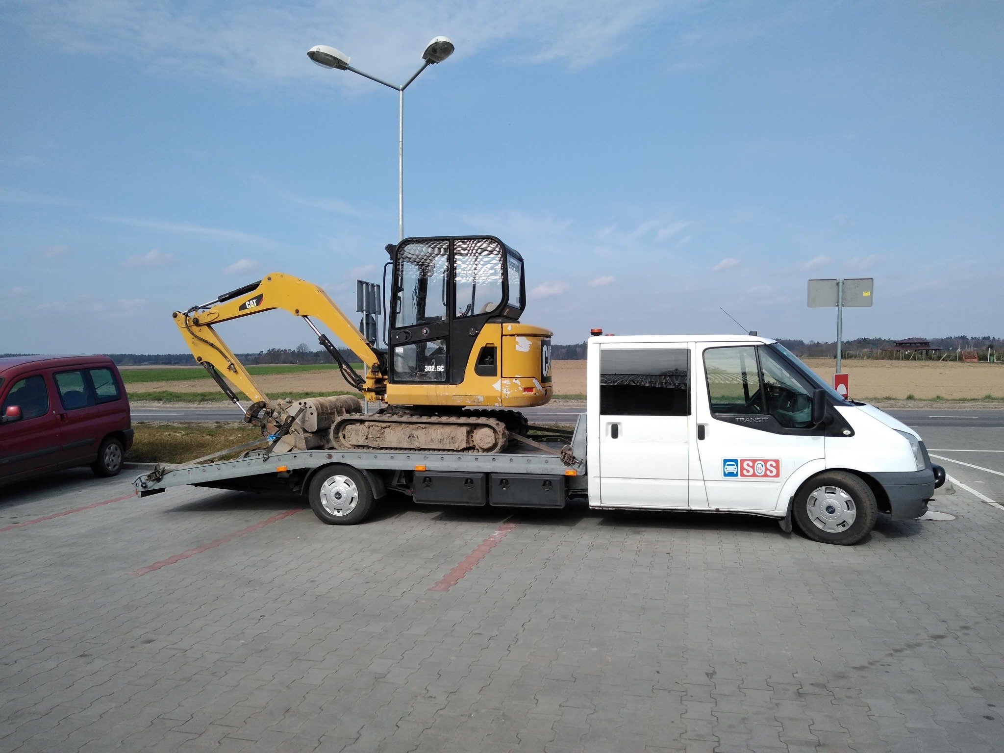 Żółta mini koparka CAT 302.5C zabezpieczona pasami na platformie białego samochodu dostawczego z napisem SOS, zaparkowanego na szarym chodniku.