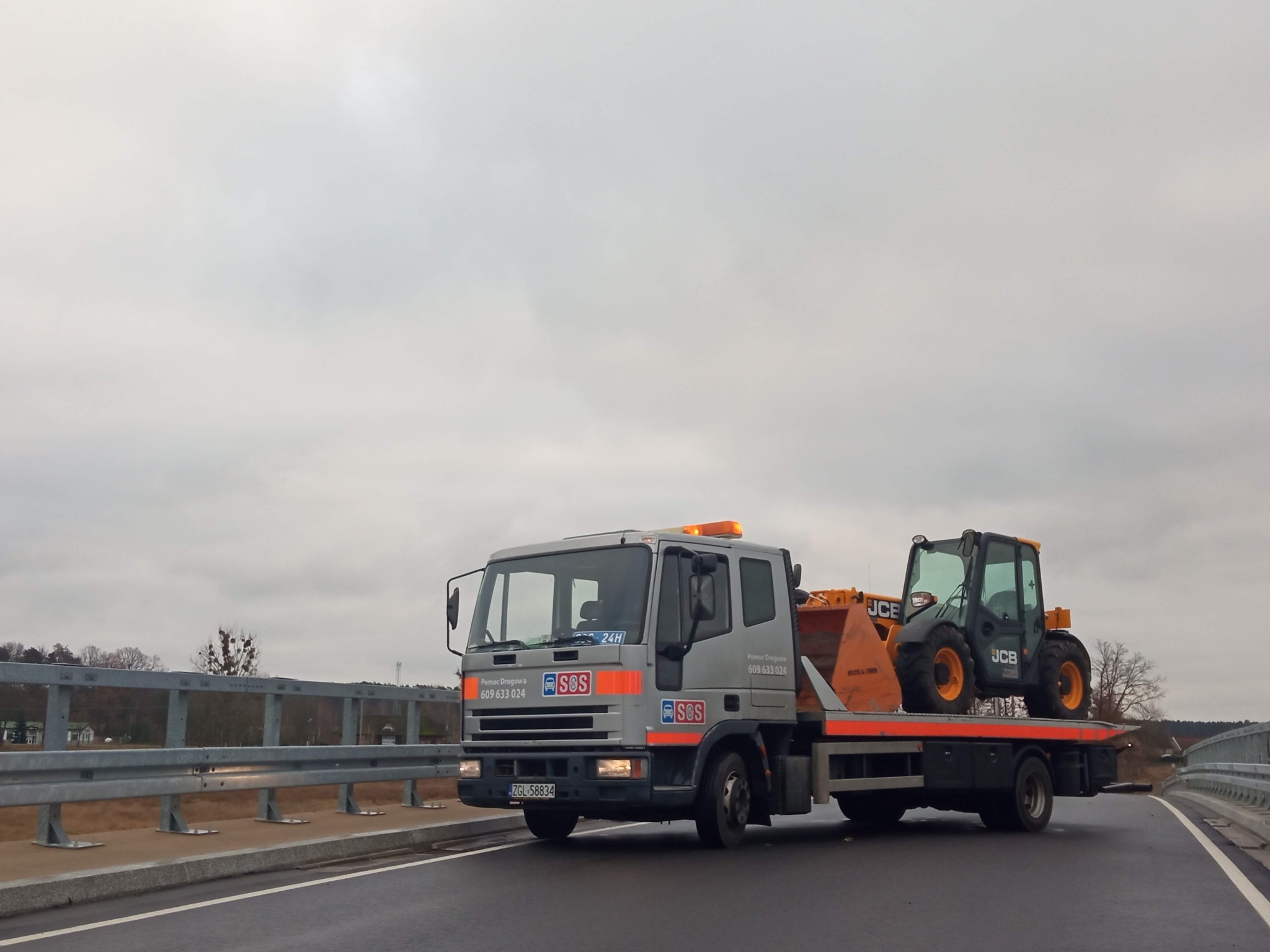Szara laweta firmy Pomoc Drogowa SOS przewozi czarno-żółtą ładowarkę teleskopową JCB na autostradzie w pochmurny dzień.