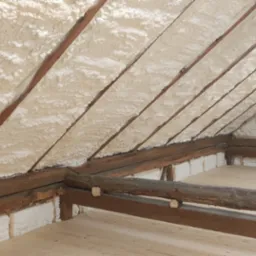 Poddasze z drewnianą konstrukcją krokwiową po natrysku pianki poliuretanowej, widoczne szczeliny wypełnione pianą, podłoga z jasnych desek, w tle stara maszyna do szycia.