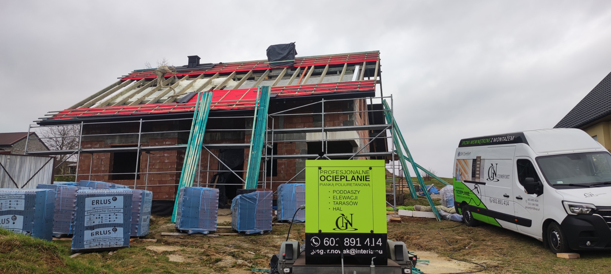 Budowa domu w trakcie prac izolacyjnych dachu: widoczne rusztowania, ułożona czerwona membrana dachowa, częściowo wypełnione pianą PUR przestrzenie między krokwiami, stosy dachówek Erlus...