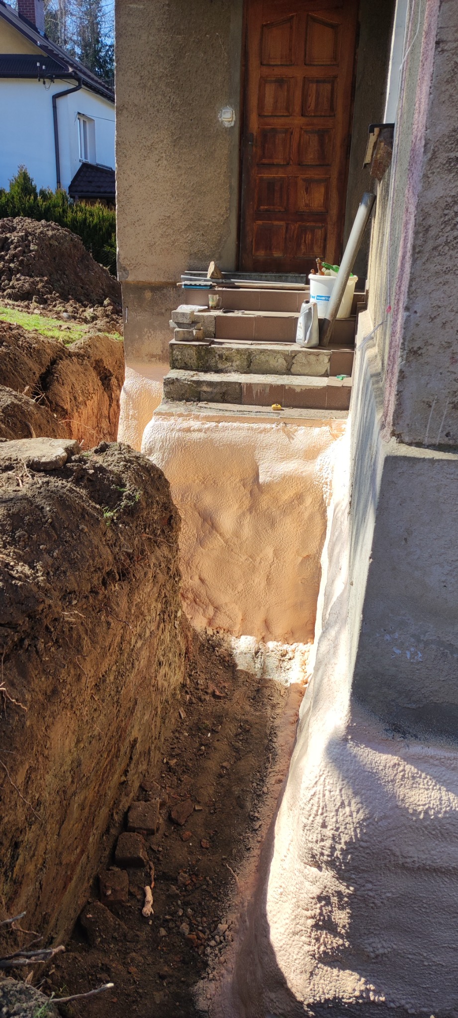 Fundament budynku z widoczną warstwą izolacyjną z pianki PUR, wykopany rów wokół fundamentu, schody wejściowe do budynku w tle.