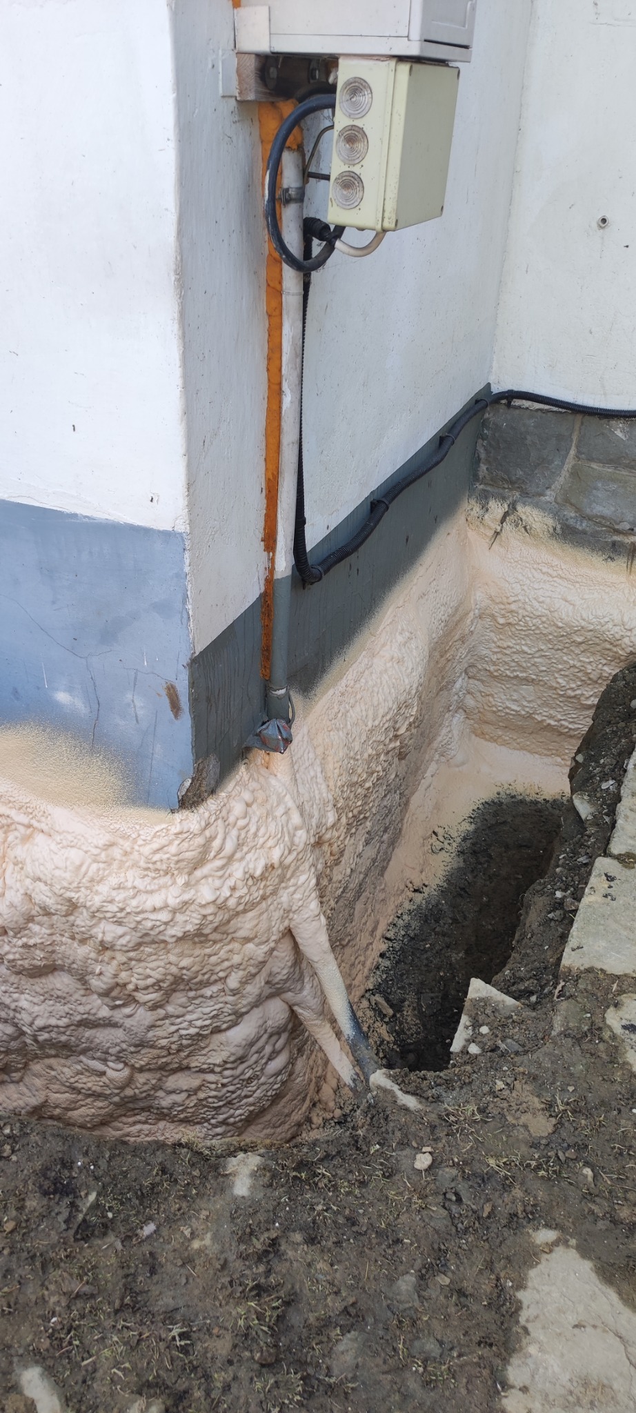 Fundament budynku w trakcie izolacji pianką poliuretanową, widoczne nierównomierne nałożenie piany i fragment wykopu z ziemią.