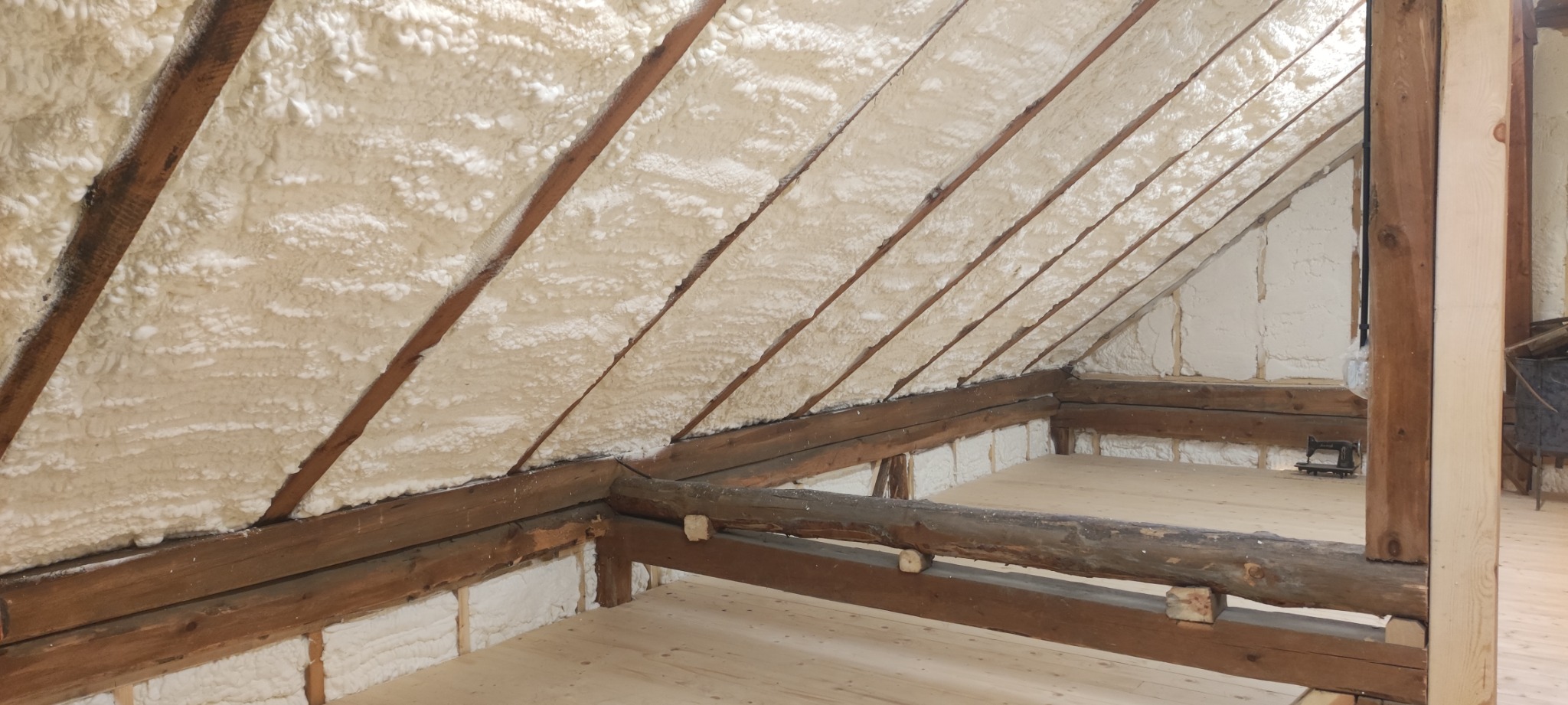 Poddasze z drewnianą konstrukcją krokwiową po natrysku pianki poliuretanowej, widoczne szczeliny wypełnione pianą, podłoga z jasnych desek, w tle stara maszyna do szycia.