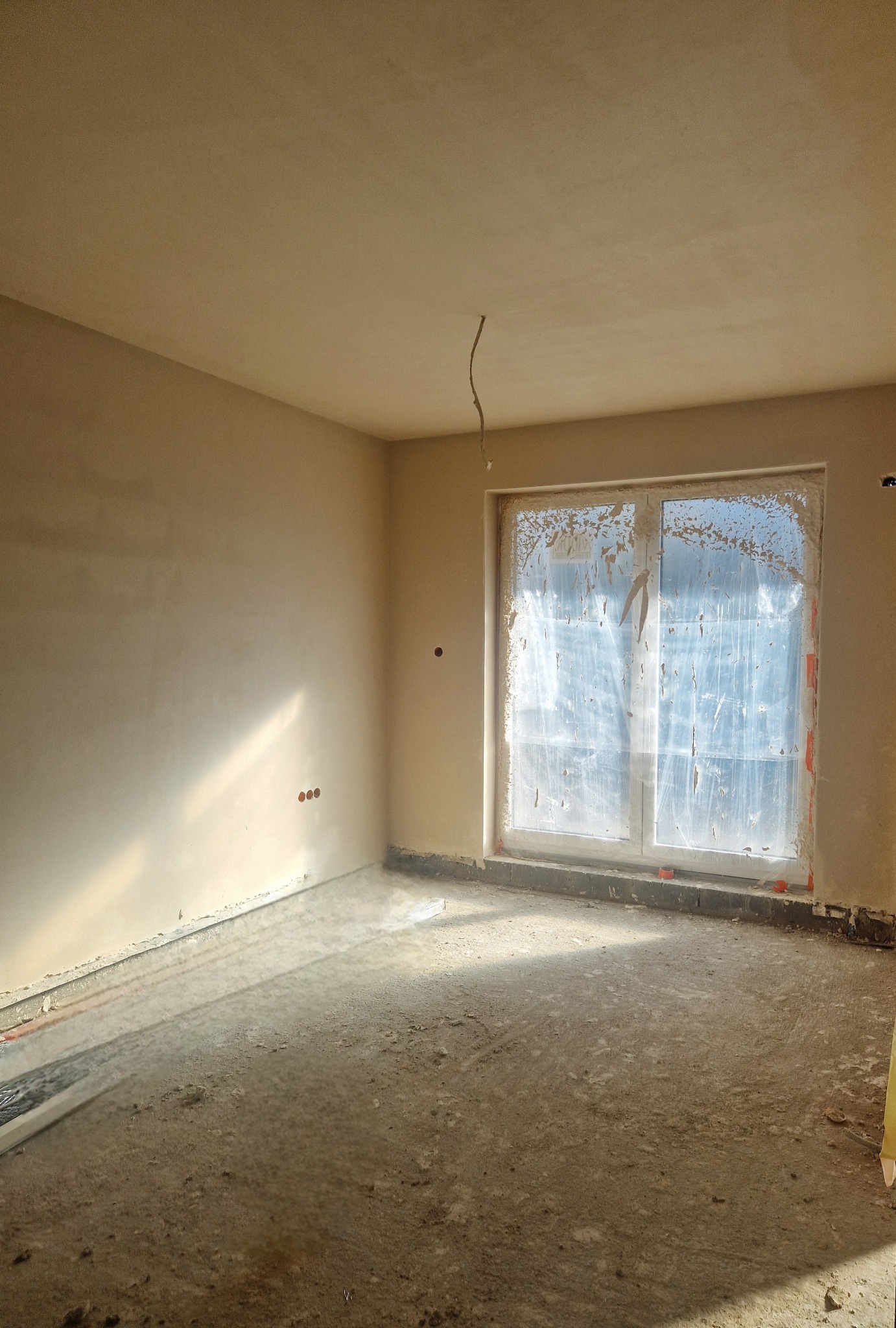 Pokój w stanie surowym po nałożeniu tynków, okno zabezpieczone folią, widoczne ślady po pracach na podłodze, świeżo otynkowane ściany w jasnym odcieniu.
