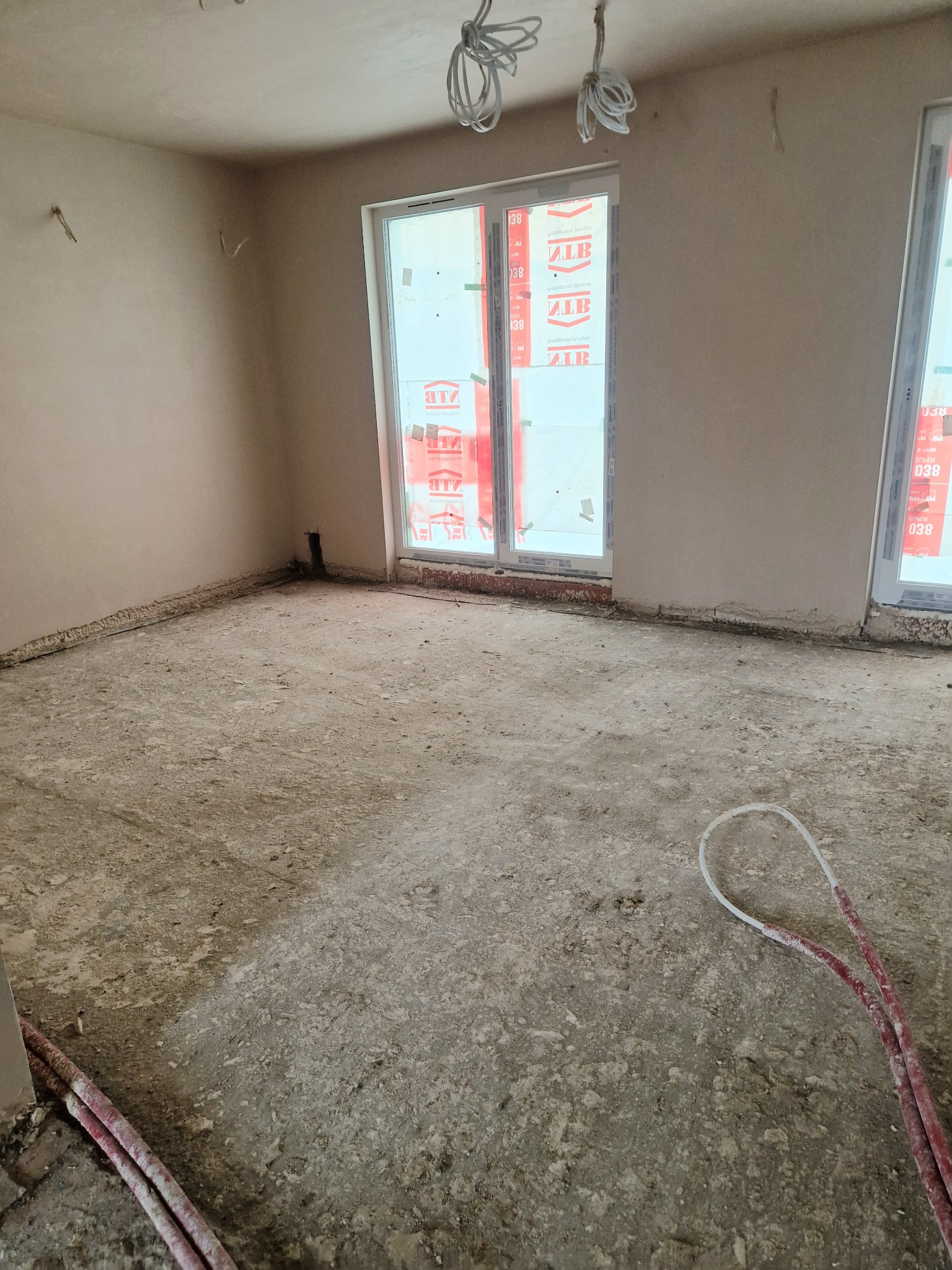 Pokój w stanie deweloperskim przed położeniem gładzi, widoczne przewody elektryczne i rury ogrzewania podłogowego. Okna zabezpieczone folią. Ściany wymagają wyrównania.