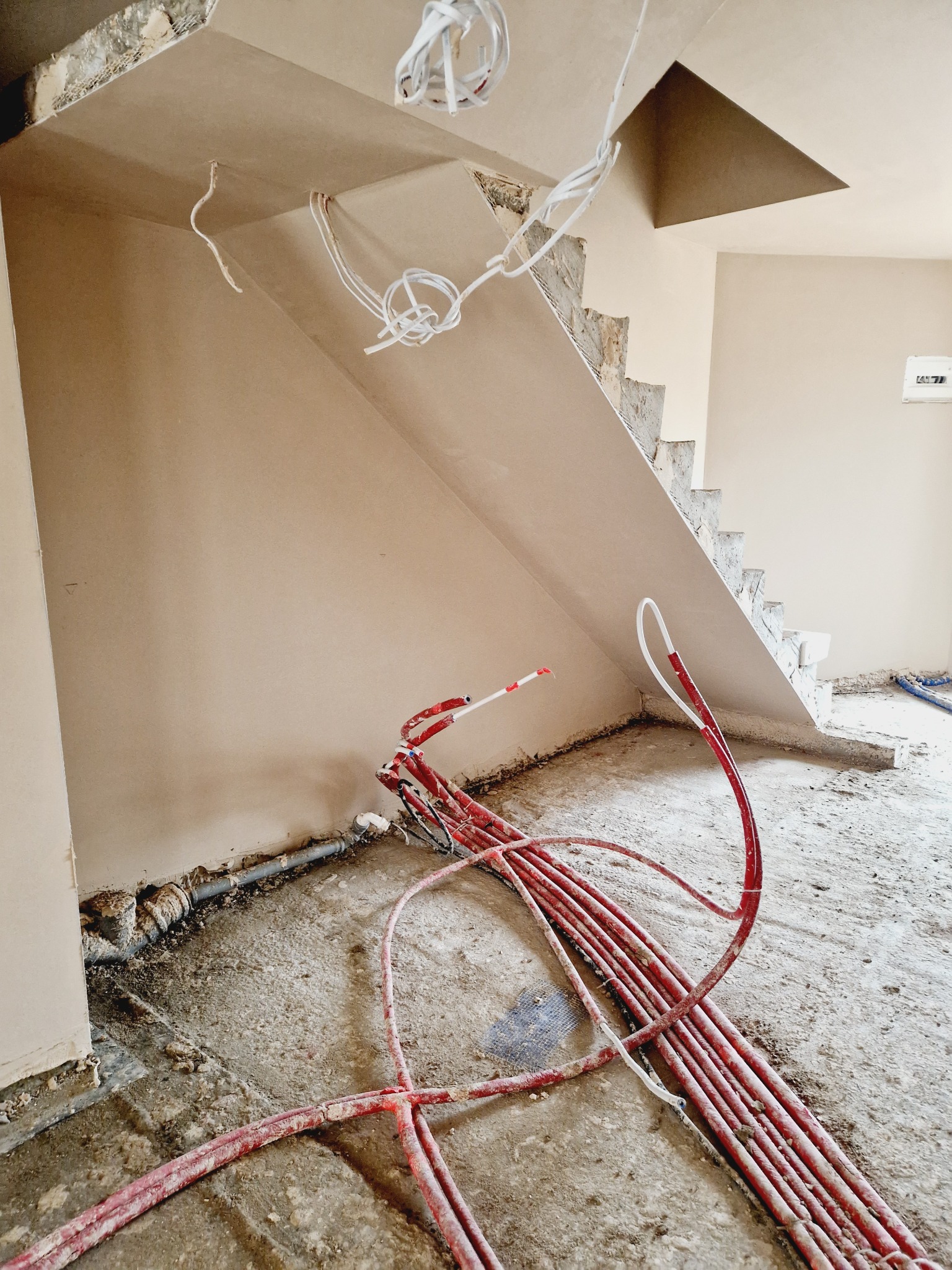 Świeżo otynkowana ściana pod schodami, z widocznymi przewodami elektrycznymi i instalacją centralnego ogrzewania w rurach koloru czerwonego, leżącymi na jeszcze nieoczyszczonej podłodze.