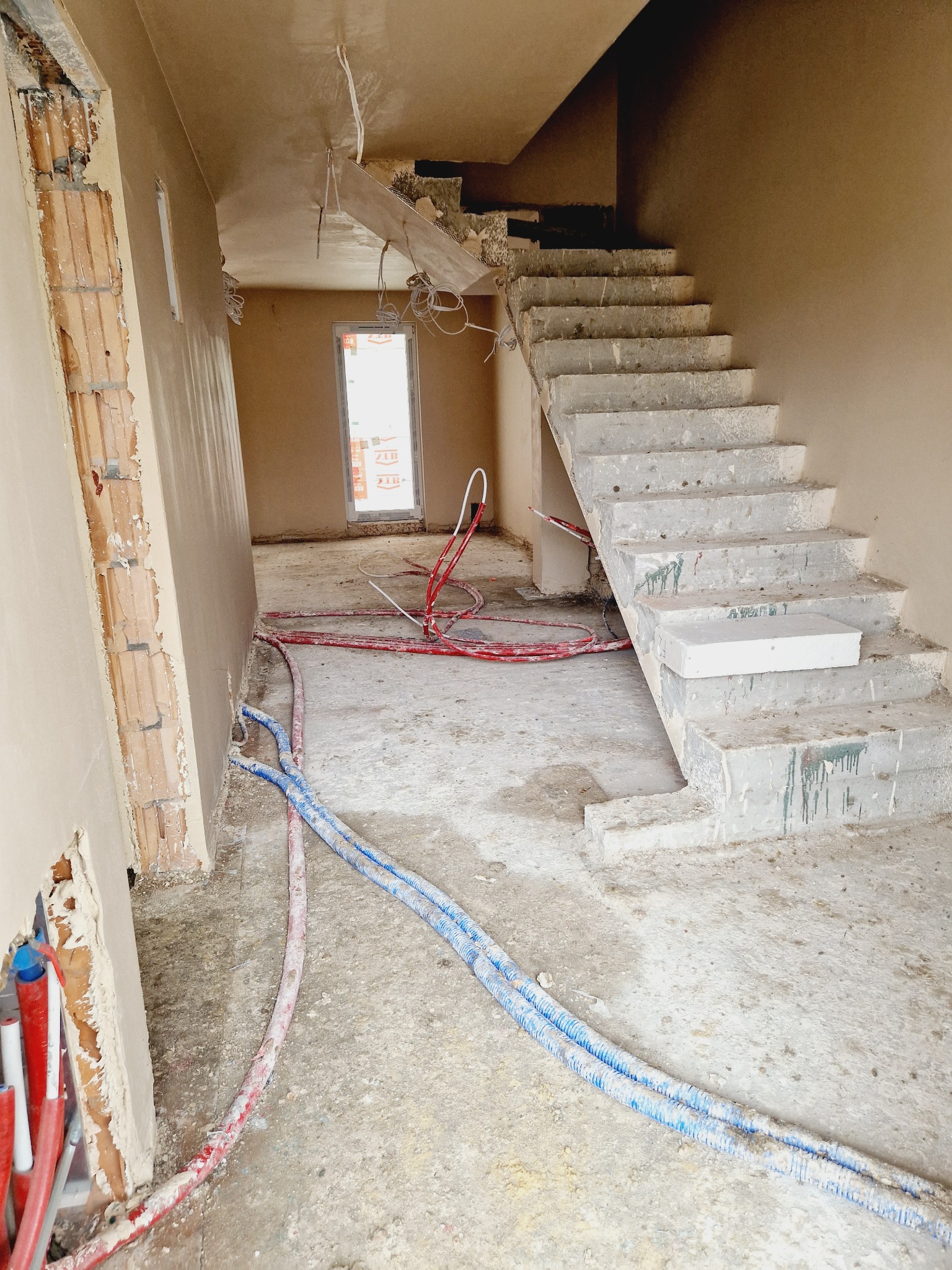 Wnętrze domu w trakcie prac wykończeniowych: ściany pokryte świeżym tynkiem, widoczne instalacje, surowe schody betonowe, okno w tle.