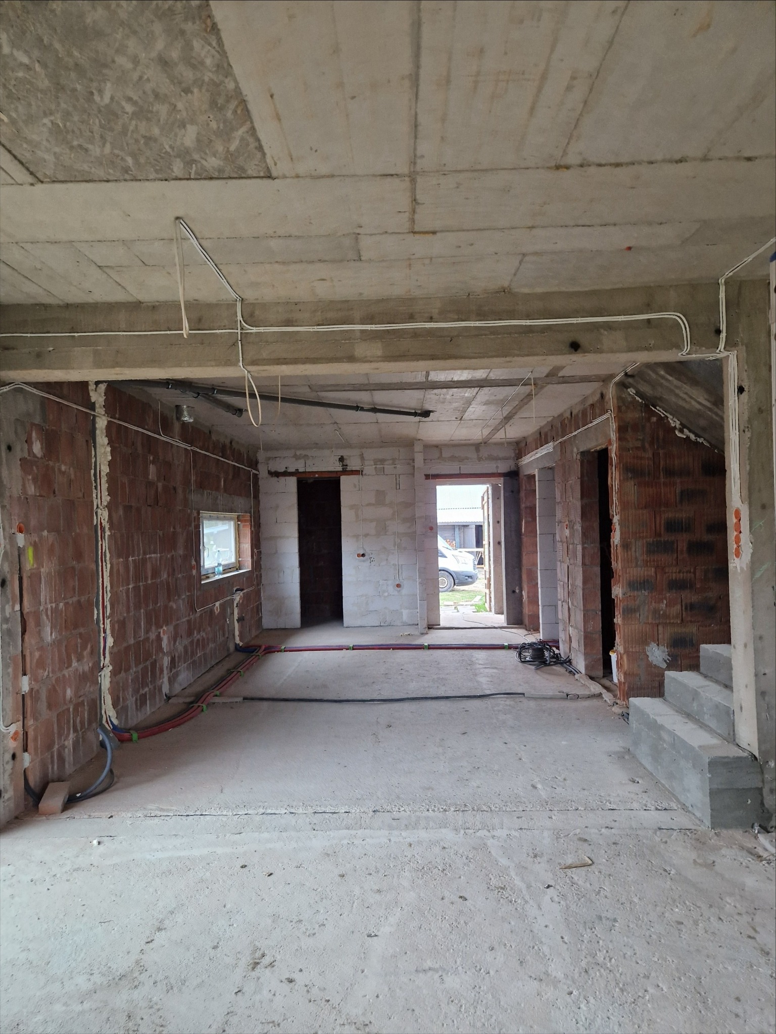 Surowe wnętrze domu w trakcie budowy z widocznymi ceglanymi ścianami, betonowym stropem i przewodami elektrycznymi. W tle widoczne okno i zarys samochodu na zewnątrz.