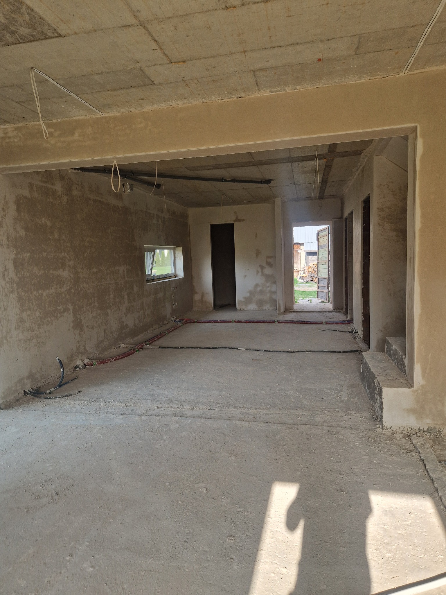 Surowe wnętrze domu w trakcie tynkowania maszynowego; widoczne przewody instalacji podłogowej i okno z widokiem na zewnątrz.