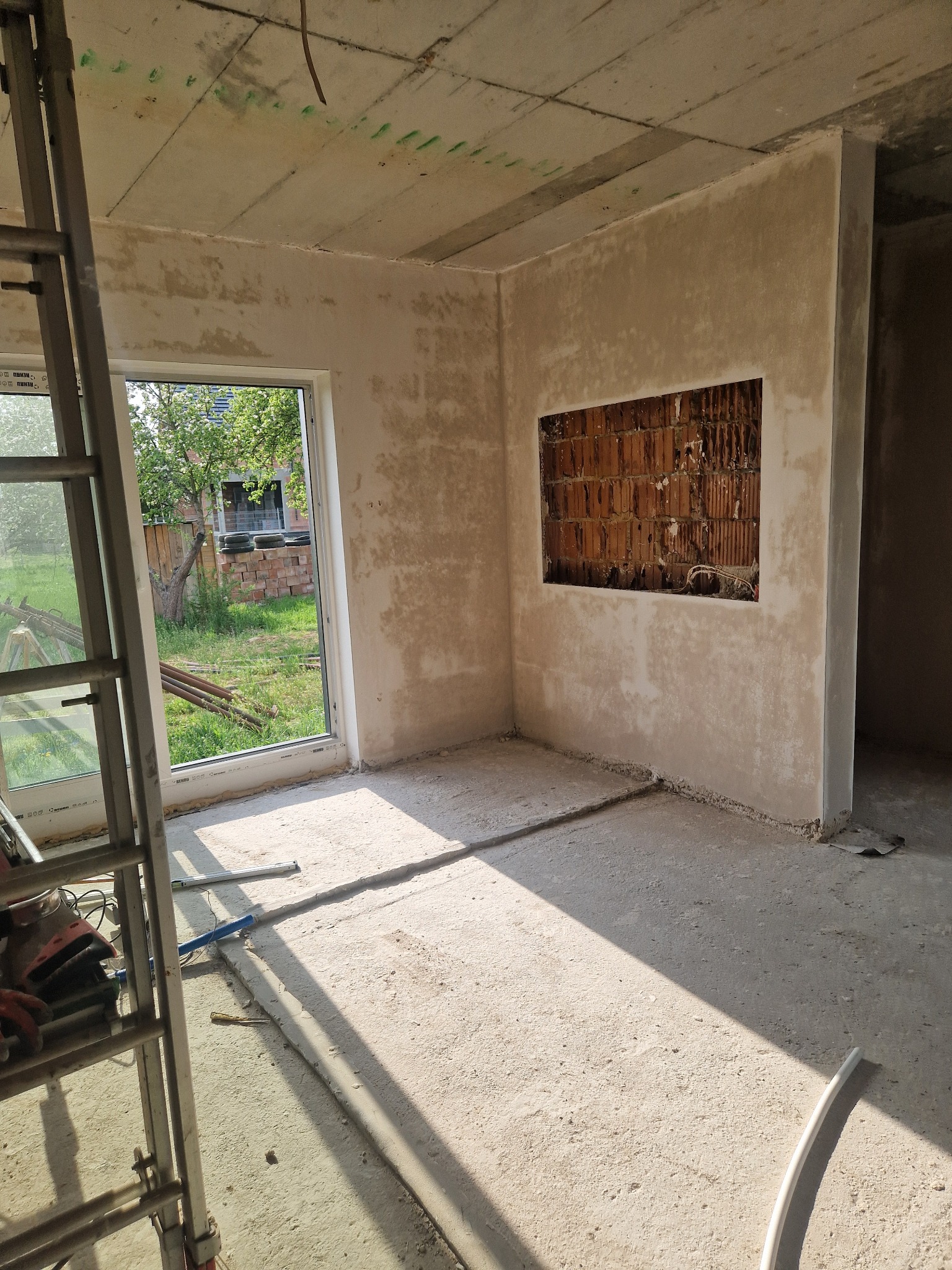 Wnętrze w trakcie prac tynkarskich: ściany częściowo pokryte tynkiem, odsłonięty fragment muru z cegły, drabina malarska przy oknie z widokiem na ogród.