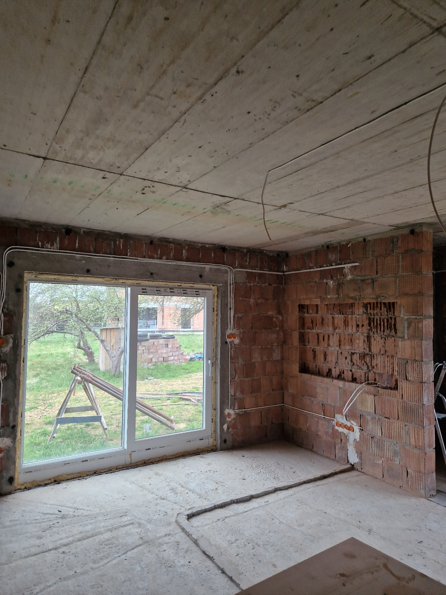 Surowy stan wnętrza z ceglanymi ścianami, betonowym sufitem i wstawionym oknem balkonowym z widokiem na ogród, widoczne instalacje elektryczne w peszlach.