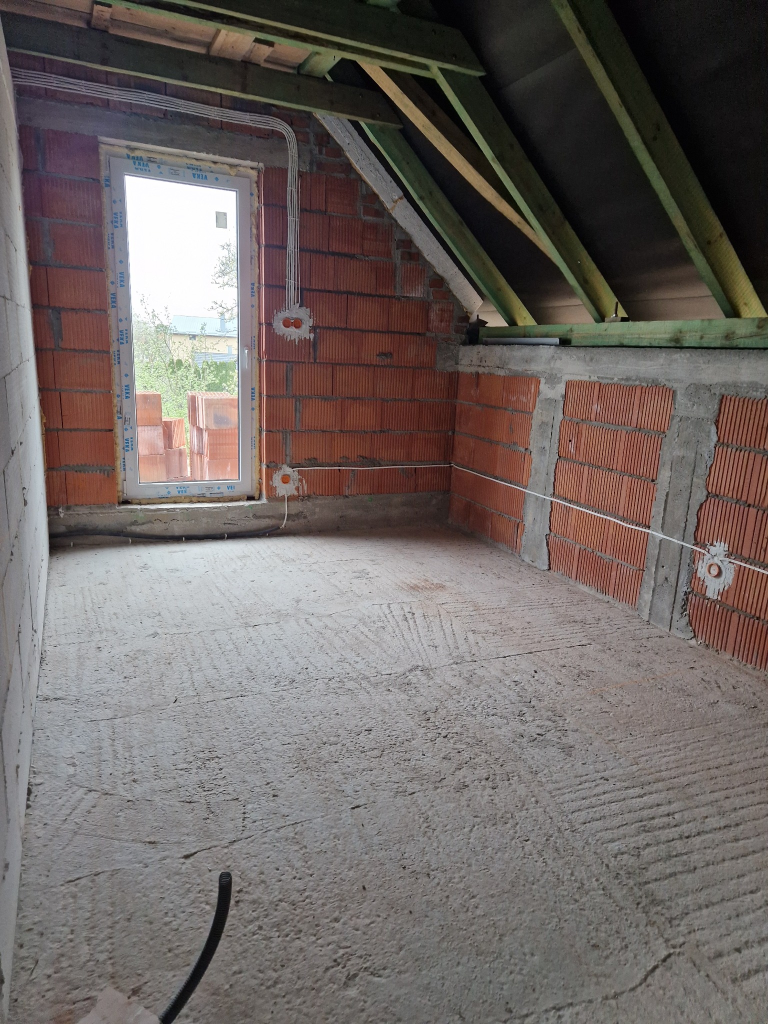 Poddasze w stanie surowym, ściany z czerwonej cegły z widocznymi przewodami elektrycznymi, okno z białymi ramami, surowy betonowy podkład, drewniana konstrukcja dachu.