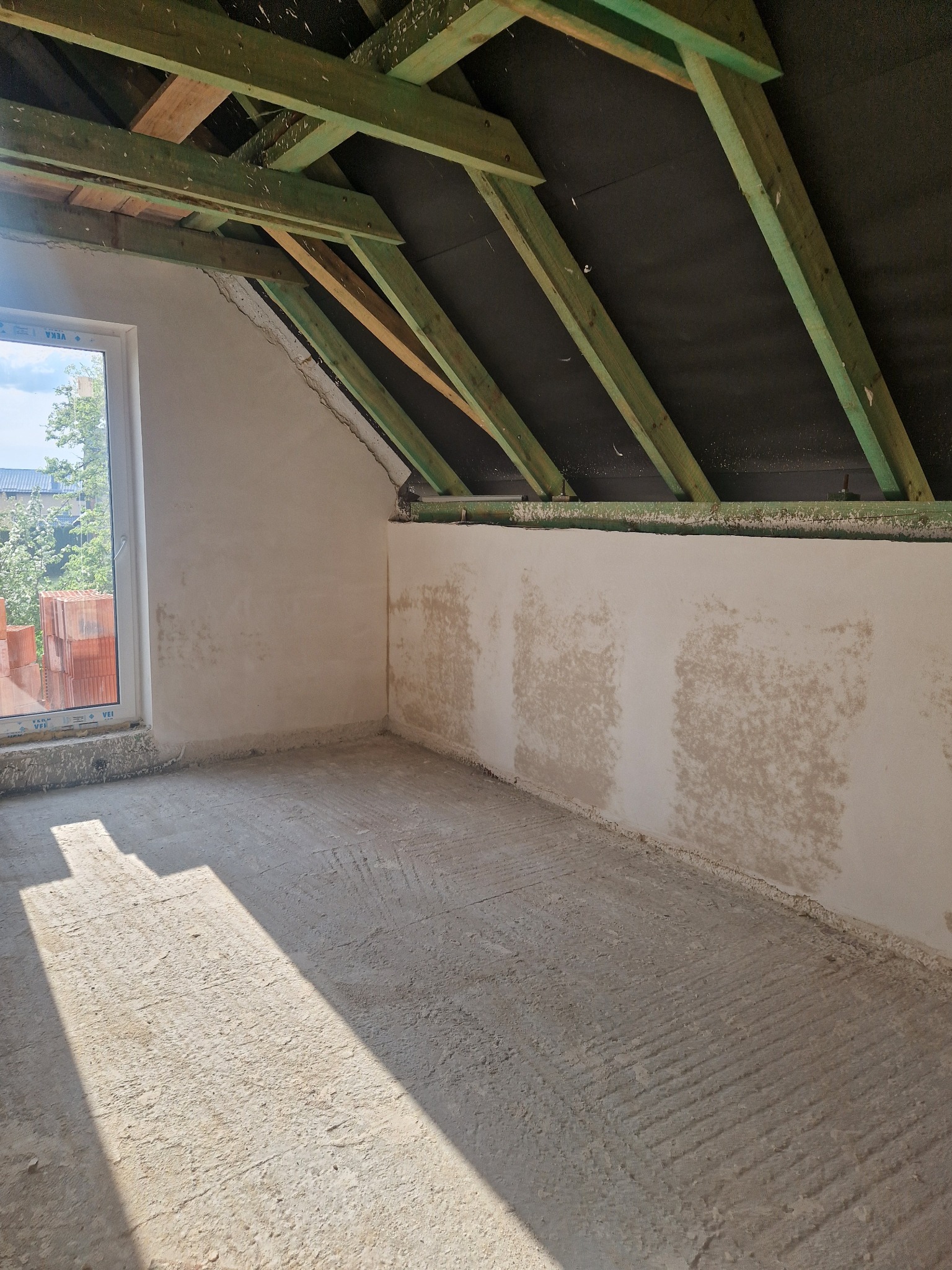 Poddasze w trakcie prac wykończeniowych: surowe tynki na ścianach, widoczne krokwie dachowe pomalowane na zielono, w oknie widok na zieleń.