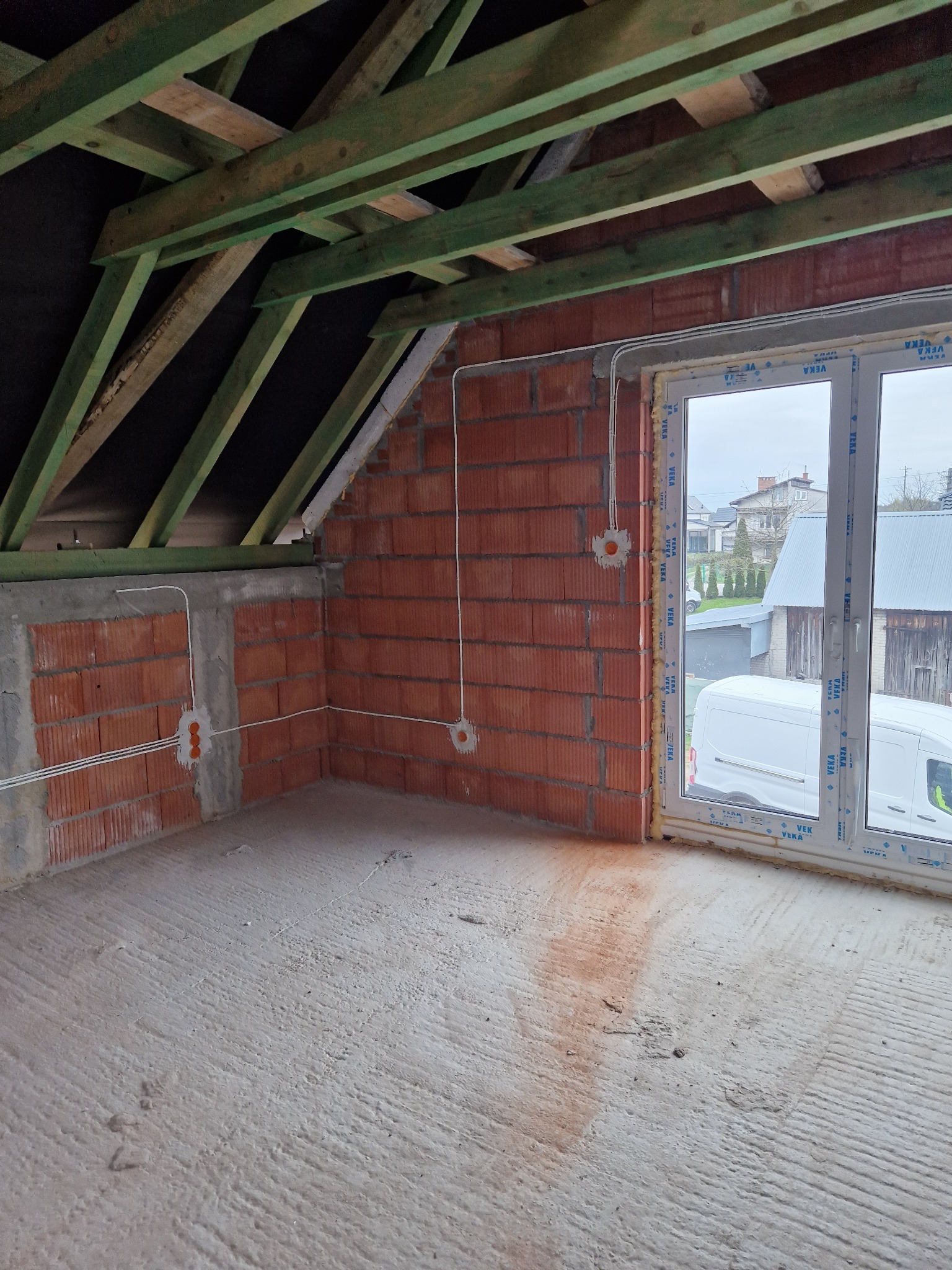 Poddasze w stanie surowym: ceglane ściany z instalacją elektryczną, drewniana konstrukcja dachu, okno PCV z widokiem na budynek gospodarczy i samochód dostawczy.