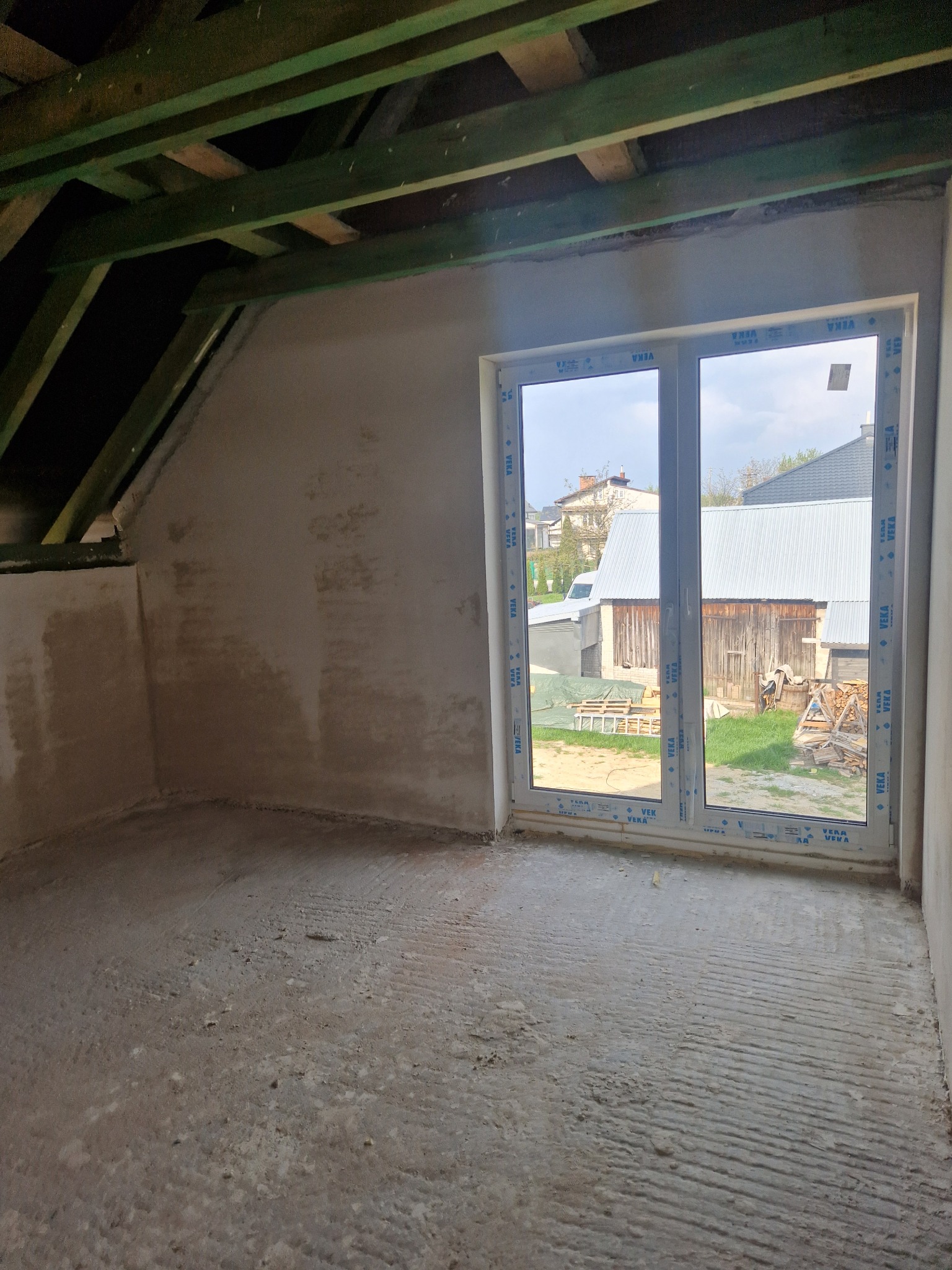 Surowe wnętrze poddasza z oknem PVC, ściany przygotowane pod prace wykończeniowe, widoczne elementy konstrukcji dachu.