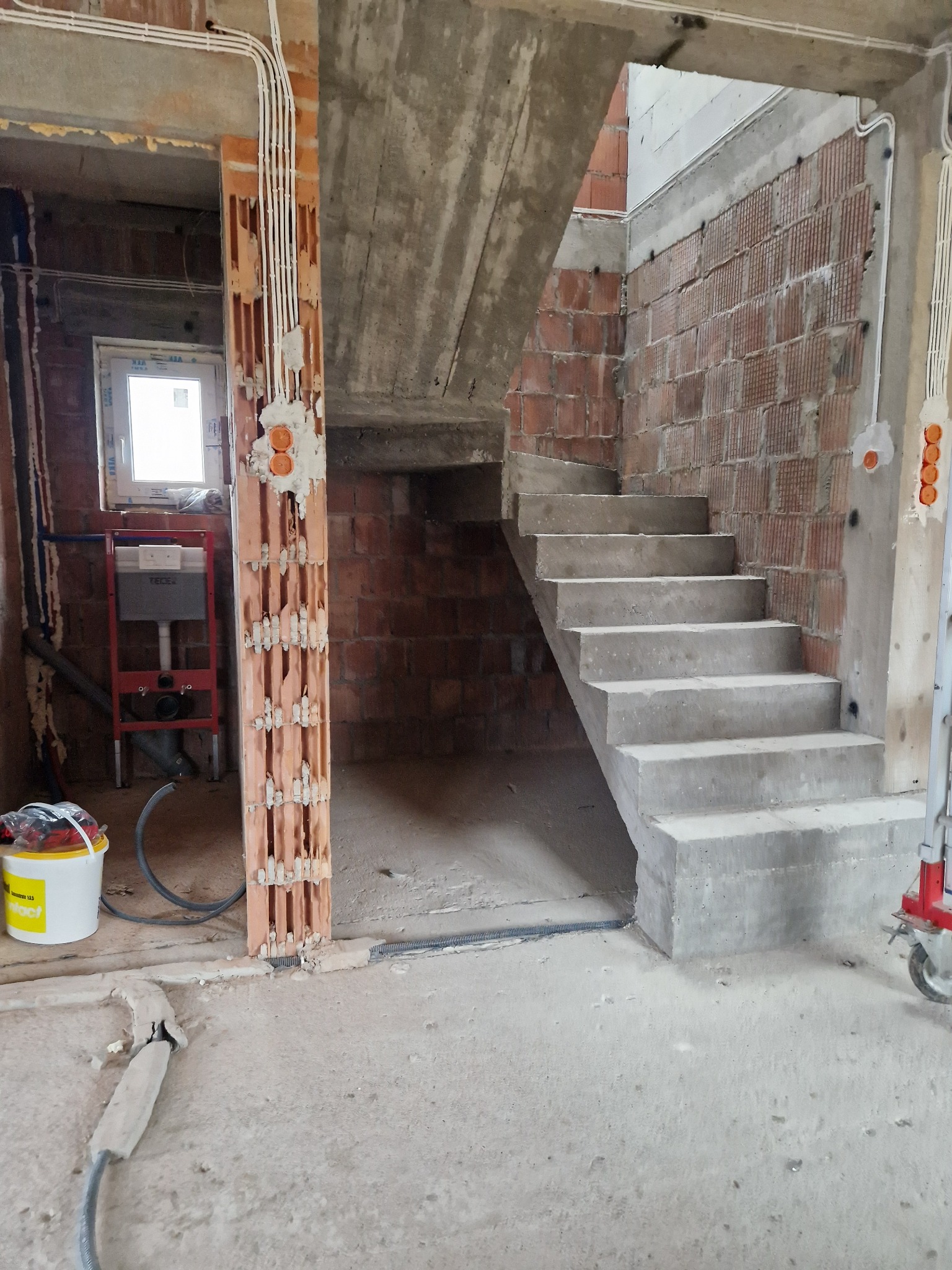 Surowe betonowe schody wewnętrzne w trakcie budowy, widoczne instalacje elektryczne i sanitarna, ceglane ściany w stanie surowym, Głogów Małopolski.