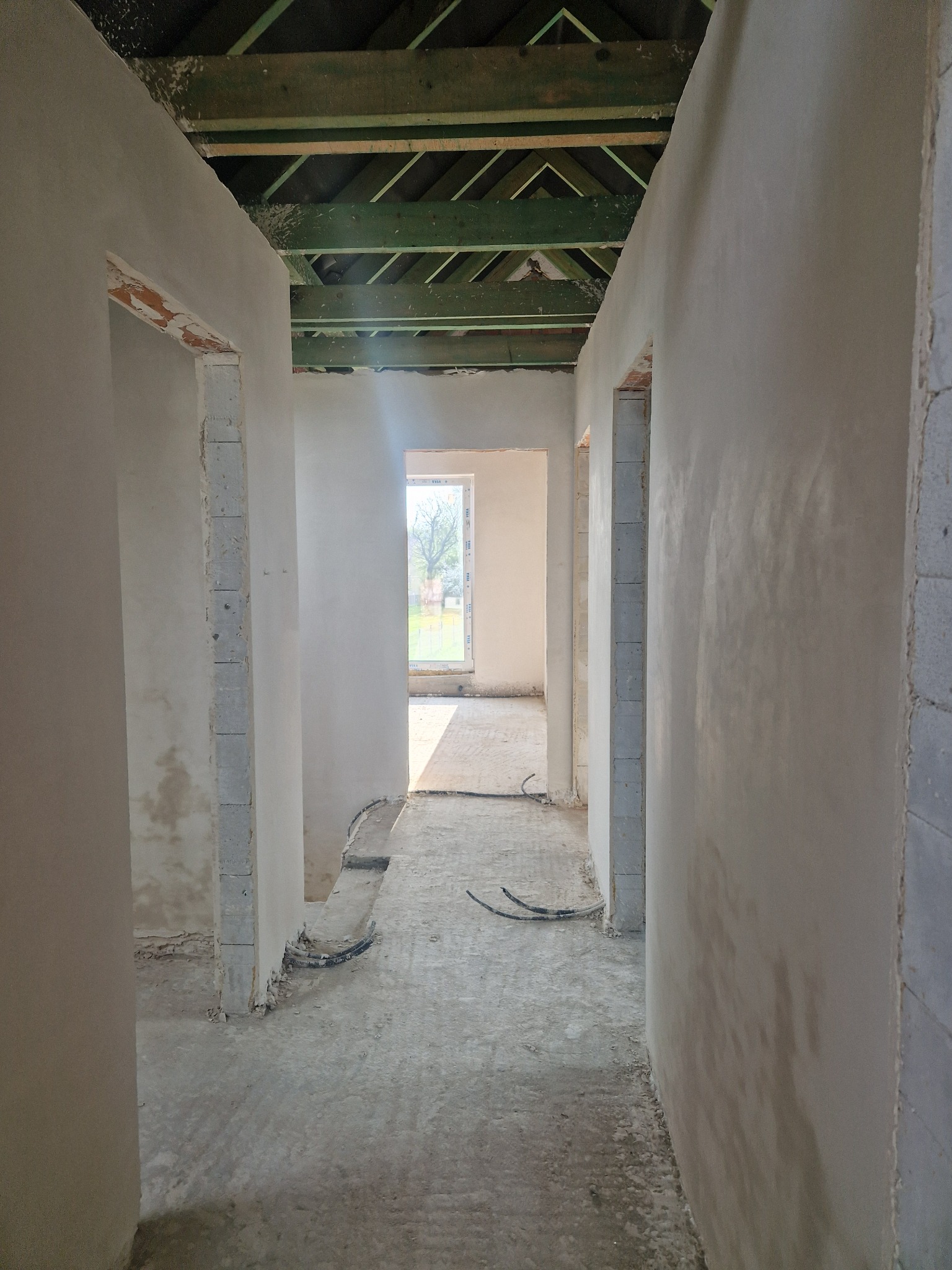 Korytarz w stanie surowym po nałożeniu tynków; widoczne ślady po pracach budowlanych na podłodze i odkryta konstrukcja dachu.