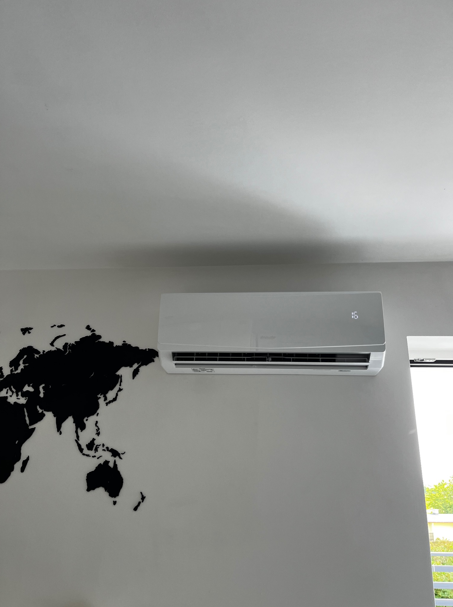 Biała klimatyzacja na ścianie z mapą świata, obok okna. Nowoczesny design, jasne wnętrze. Widoczna temperatura 16 stopni.