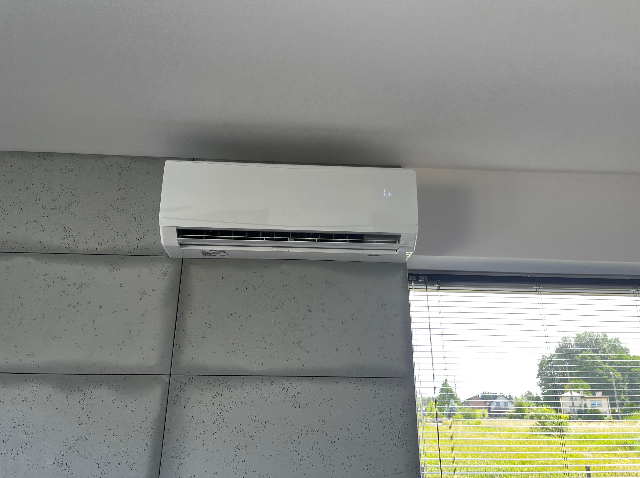 Biała klimatyzacja ścienna w nowoczesnym wnętrzu z betonową ścianą i widokiem na zieleń przez okno z żaluzjami. Minimalistyczny design.