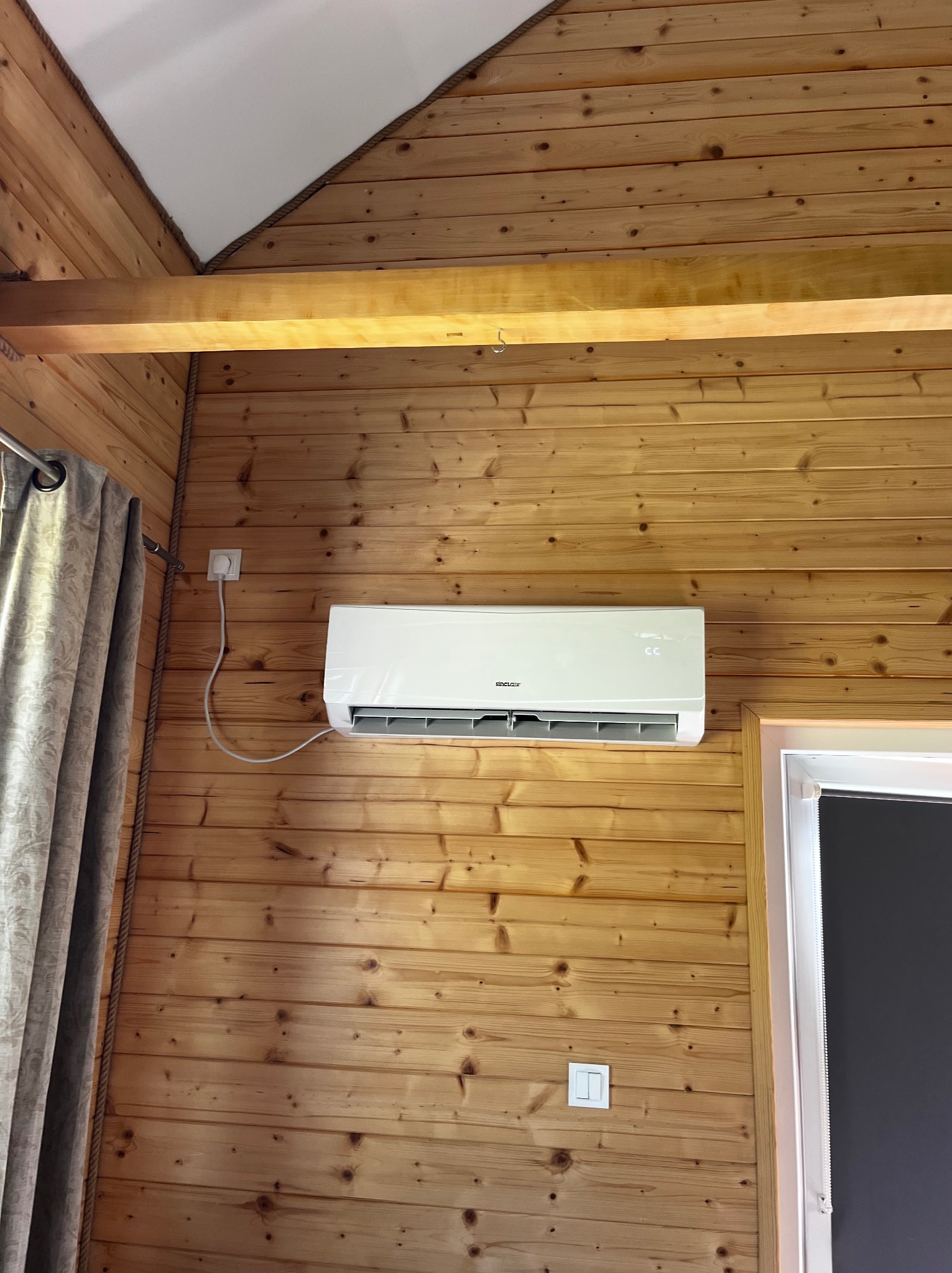 Biała klimatyzacja Sinclair zamontowana na ścianie z boazerii w drewnianym wnętrzu. Widoczny fragment okna i zasłony. Kabel zasilający podłączony do gniazdka.